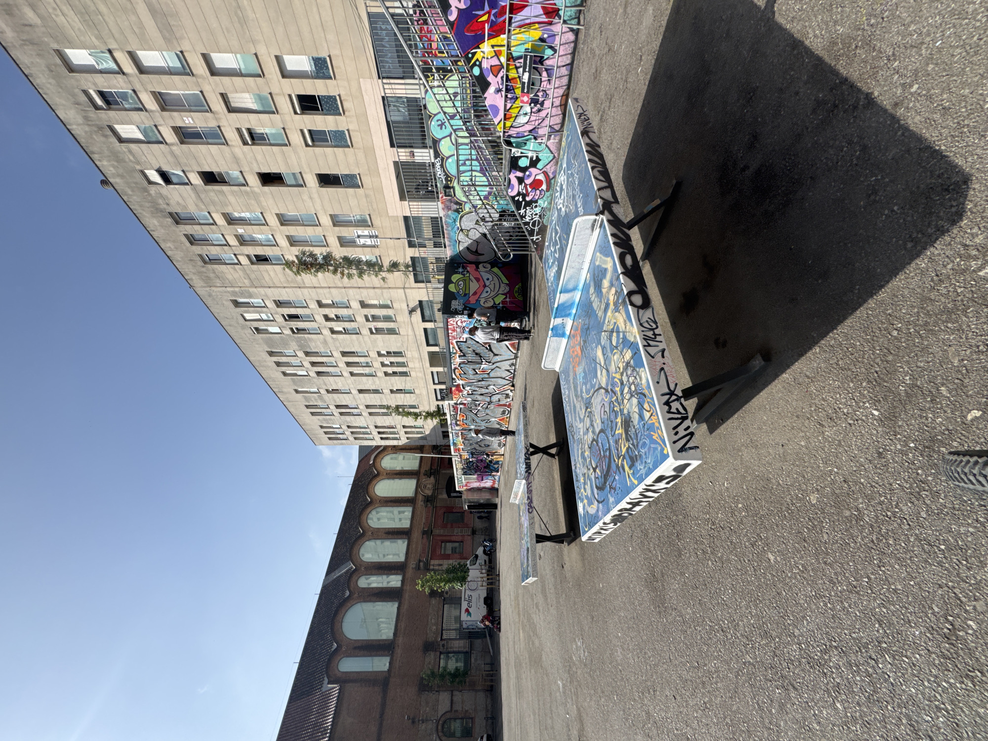 The image, captured from a low angle and rotated 90 degrees clockwise, depicts an outdoor urban setting featuring prominent architecture and vibrant graffiti art. To the viewer's left (when oriented correctly), a tall, light-grey concrete building with multiple rows of rectangular windows stands against a clear blue sky. To the right, a darker, brick-like building with distinctive arched windows on its upper facade provides a contrasting architectural style.
In the center, an area dedicated to urban art displays extensive, colorful graffiti covering a wall and metal railings. Several large, rectangular canvases or panels, propped on black metal stands, are heavily adorned with intricate graffiti. The most prominent panel, predominantly blue with black and white tags, clearly shows the letters "N.V.X.R." in white, along with "S.T.N.R.K" and "Z.E.R.M." on its edges.
The ground is a paved asphalt surface, across which long, sharp shadows are cast, indicating bright sunlight, likely in the morning or late afternoon. A large, dark shadow dominates the foreground on the right. Two individuals are visible near the graffiti wall, one standing and another crouching, possibly artists or observers. A white van with an "Elis" logo and a red scooter are parked nearby, and a bicycle tire is partially visible in the bottom right corner. The scene conveys a dynamic, artistic urban atmosphere, suggestive of a street art exhibition or a creative hub in a city like Barcelona.