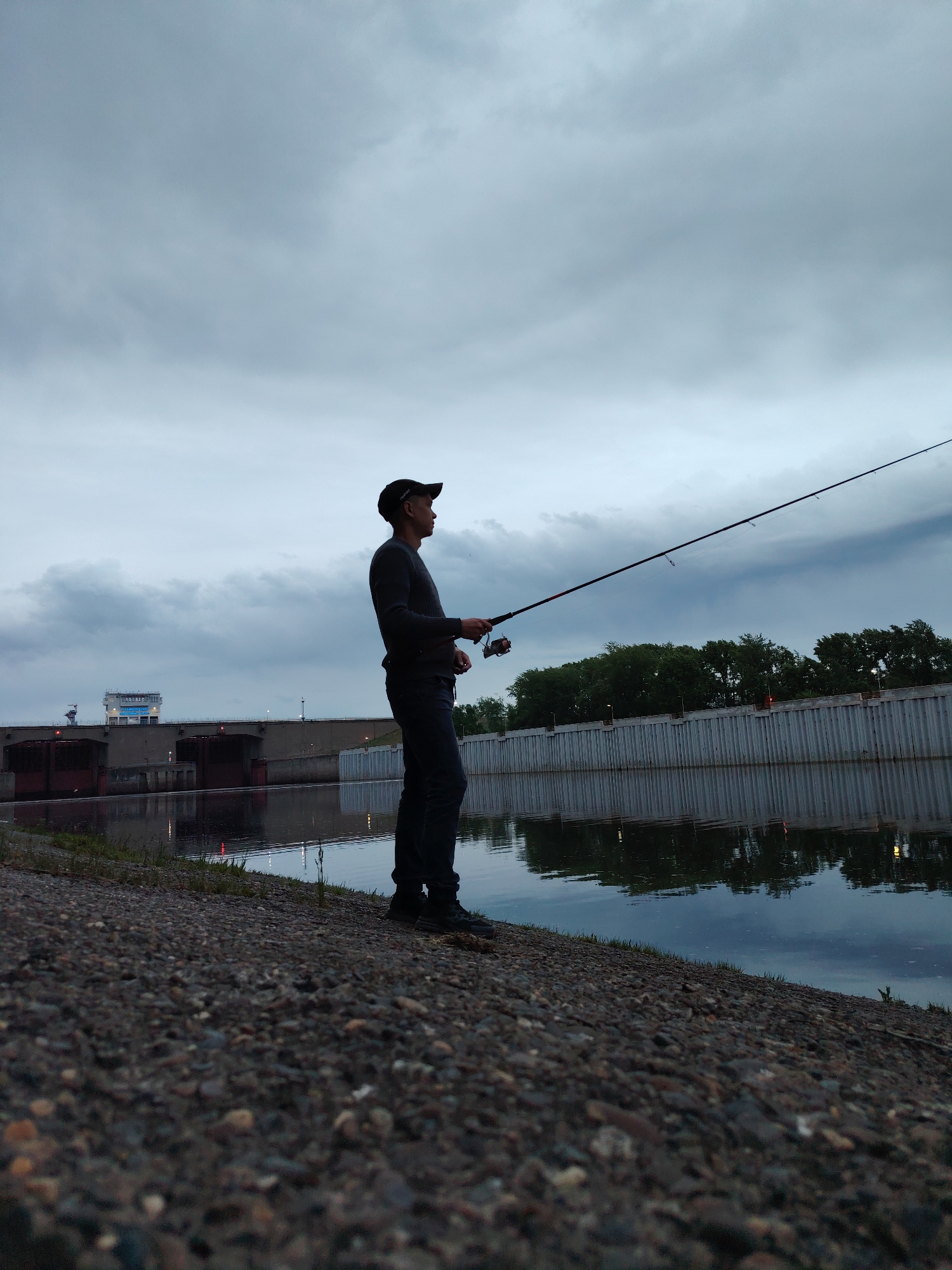 The image depicts a man fishing on the bank of a river or canal in what appears to be Chaikovsky, Russia. The man, wearing a dark long-sleeved shirt, dark pants, and a cap, is holding a fishing rod extended over the water. A dam structure is visible in the background to the left. The water is still, reflecting the overcast sky and the embankment walls on the opposite bank. The bank where the man is standing is composed of small rocks and sparse vegetation. The sky is cloudy, suggesting either early morning or evening light.