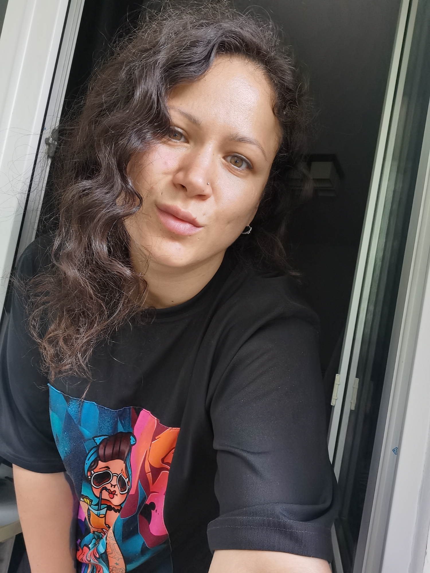 A young woman with curly dark brown hair and light brown eyes is looking at the camera. She is wearing a black t-shirt with a colorful graphic print of a woman with sunglasses. She has a nose ring and a small hoop earring in her left ear. The background appears to be an open doorway or window, with a white frame and dark interior. The lighting suggests it is daytime, with light coming from the left side of the frame. The specific location in Chaikovsky, Russia, is not discernible from the image.