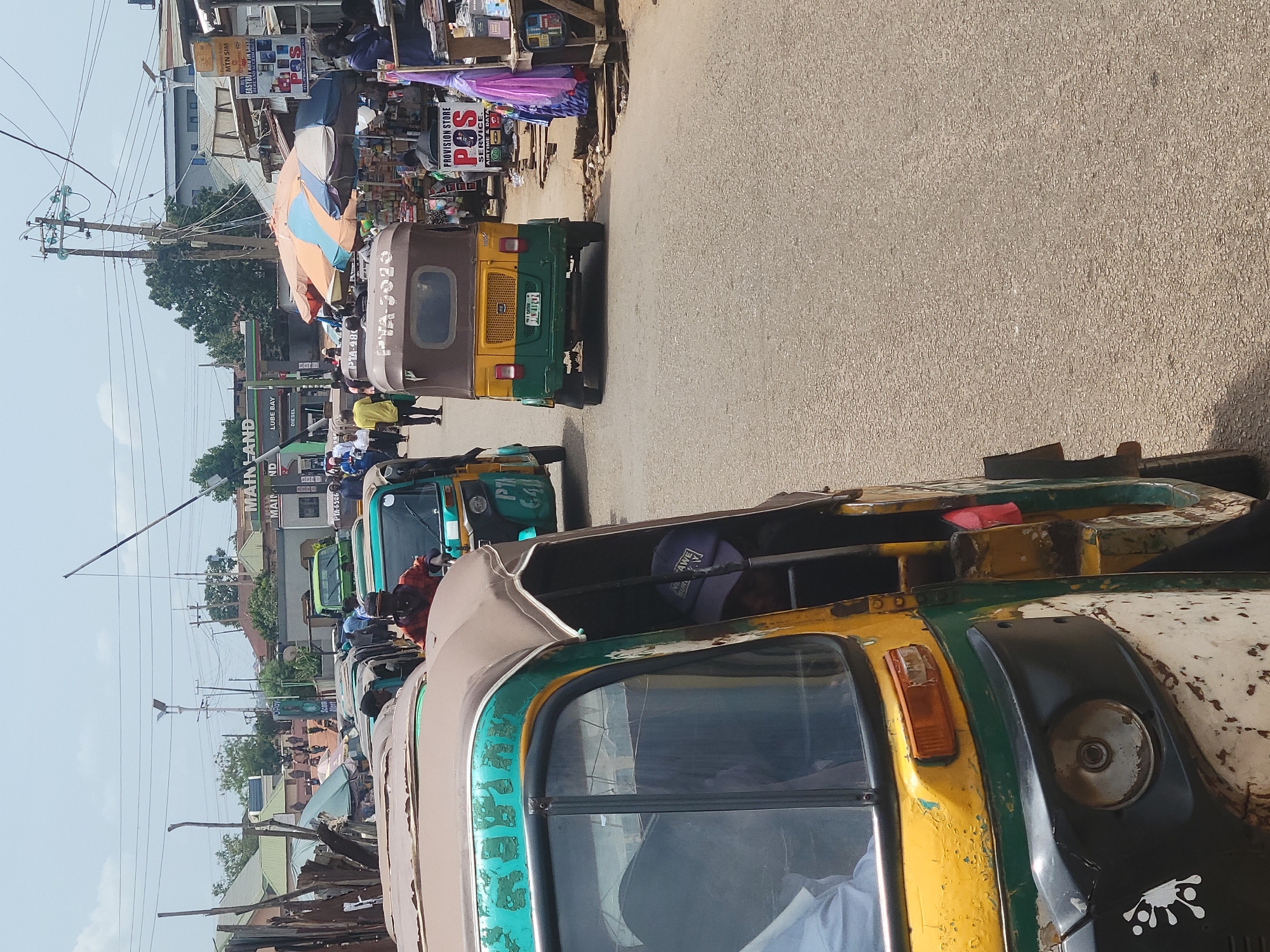 The image depicts a street in Jos, Nigeria, crowded with auto-rickshaws, locally known as 'Keke Napep'. The foreground features a dilapidated yellow and green Keke Napep with a visible headrest, occupied by a driver wearing a purple cap. The middle ground presents a line of Keke Napeps in shades of green and brown. Signs for 'Mainland' and 'Lube Bay' are evident among the buildings. Pedestrians navigate alongside the vehicles. Overhead power lines crisscross the sky. A 'Provision Store' sign with the term "POS SERVICE" is visible amidst street vendor stalls. The license plate 'PTA-6526' is visible on one of the auto-rickshaws.