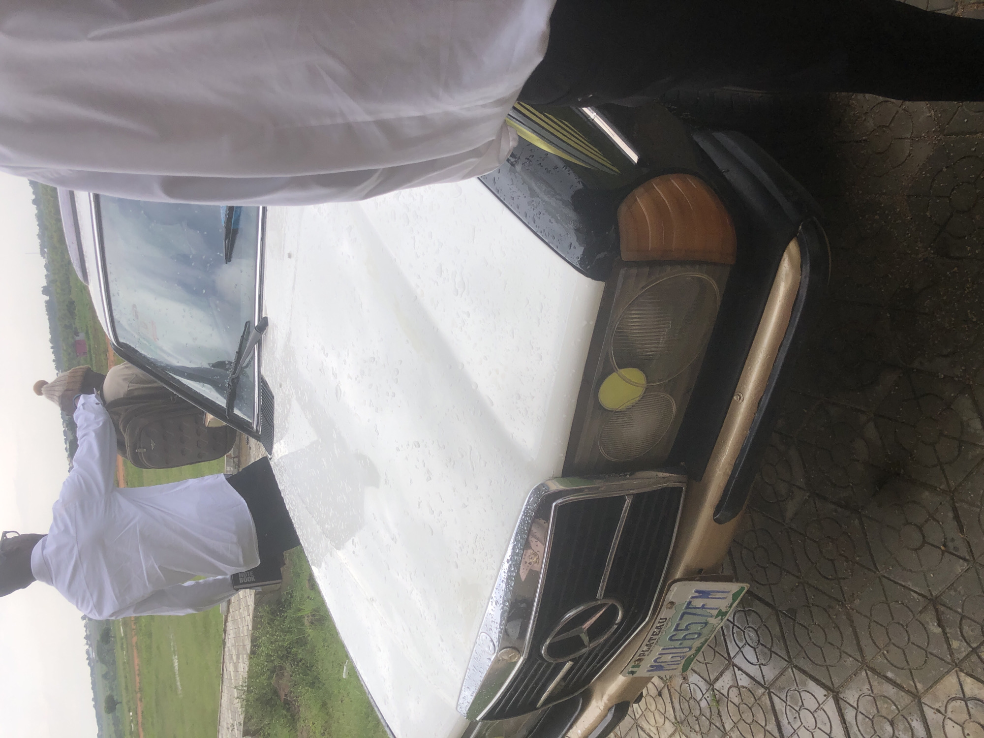 This media file depicts two individuals and a white Mercedes-Benz car, viewed from a slightly elevated, tilted perspective. The scene appears to be outdoors, possibly in a paved area with a grassy field and distant trees. The weather seems to be rainy, as indicated by water droplets on the car's hood and a wet appearance on the ground.
One person, wearing a white shirt, is visible near the car's open driver-side door, facing away from the camera and towards the grassy field. Another person, partially obscured by a white garment, is standing near the car's hood.
The car is a classic Mercedes-Benz model, identifiable by its distinct grille and headlights. A license plate is visible on the front of the car, with the text "MGH-657FM" and "PLATEAU". The overall impression is that of a casual outdoor setting, potentially in Nigeria given the "PLATEAU" text on the license plate, which could refer to Plateau State. The overcast sky and wet conditions suggest it might be a cloudy or rainy day.