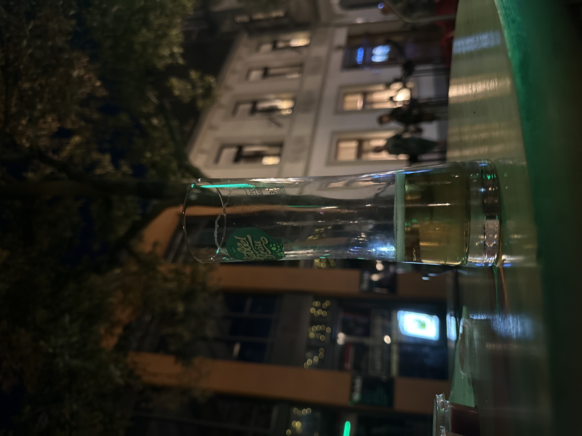 The image shows a glass of beer on a table in front of a building at night. The glass has a green label with a logo that reads "Gubbel Bräu". The building appears to be a restaurant or bar, with lights on in the windows. There are a few people in the background, but they are out of focus. The scene is dark, with some ambient light from the street and the building. It is possible this is in Krefeld, Germany, as the beer label could be a local brewery.
