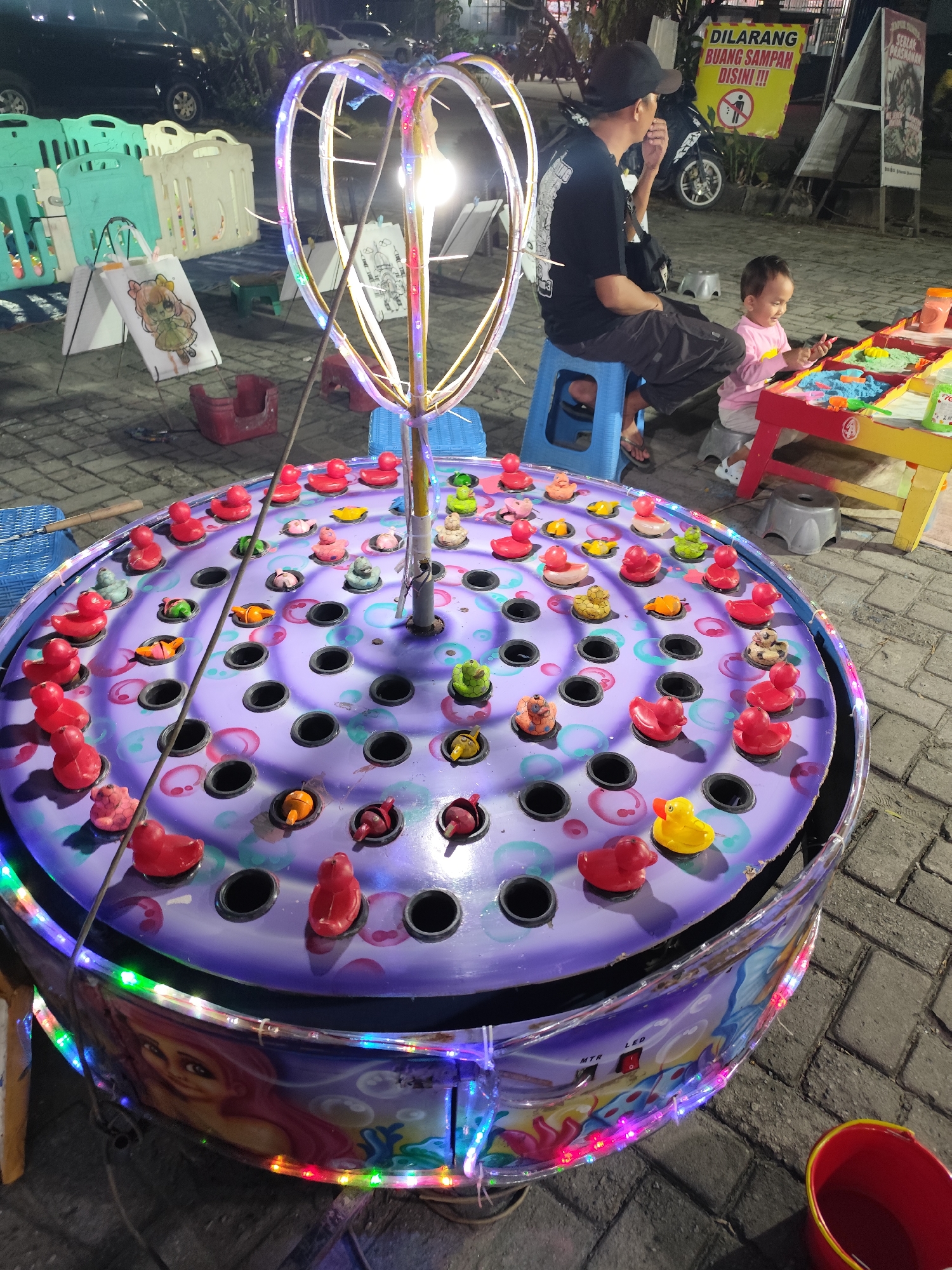 The image shows an outdoor amusement or carnival game at night. The main subject is a large, circular game with a purple top, featuring many small rubber ducks of various colors, including red, yellow, and green, floating in what appears to be water. The game also has several black holes scattered across its surface. A tall, illuminated, heart-shaped structure rises from the center of the game, adorned with colorful LED lights. The base of the game is decorated with a colorful illustration of a mermaid.
In the background, a man and a young child are visible. The man, wearing a black t-shirt and a black cap, is sitting on a blue stool. The child, dressed in a pink long-sleeved shirt, is sitting on a stool and appears to be playing with colorful sand or modeling clay on a red and yellow table. Other game equipment, like plastic barriers and small stools, are also present. The ground is paved with interlocking bricks, and the scene is illuminated by artificial lighting, suggesting it is nighttime.
A yellow sign with red text is mounted on a pole behind the man, displaying a warning about littering. Another sign with Indonesian text is partially visible to the right. A red bucket is placed on the ground in the lower right corner. The overall atmosphere seems to be one of casual entertainment, likely at a local fair or marketplace.