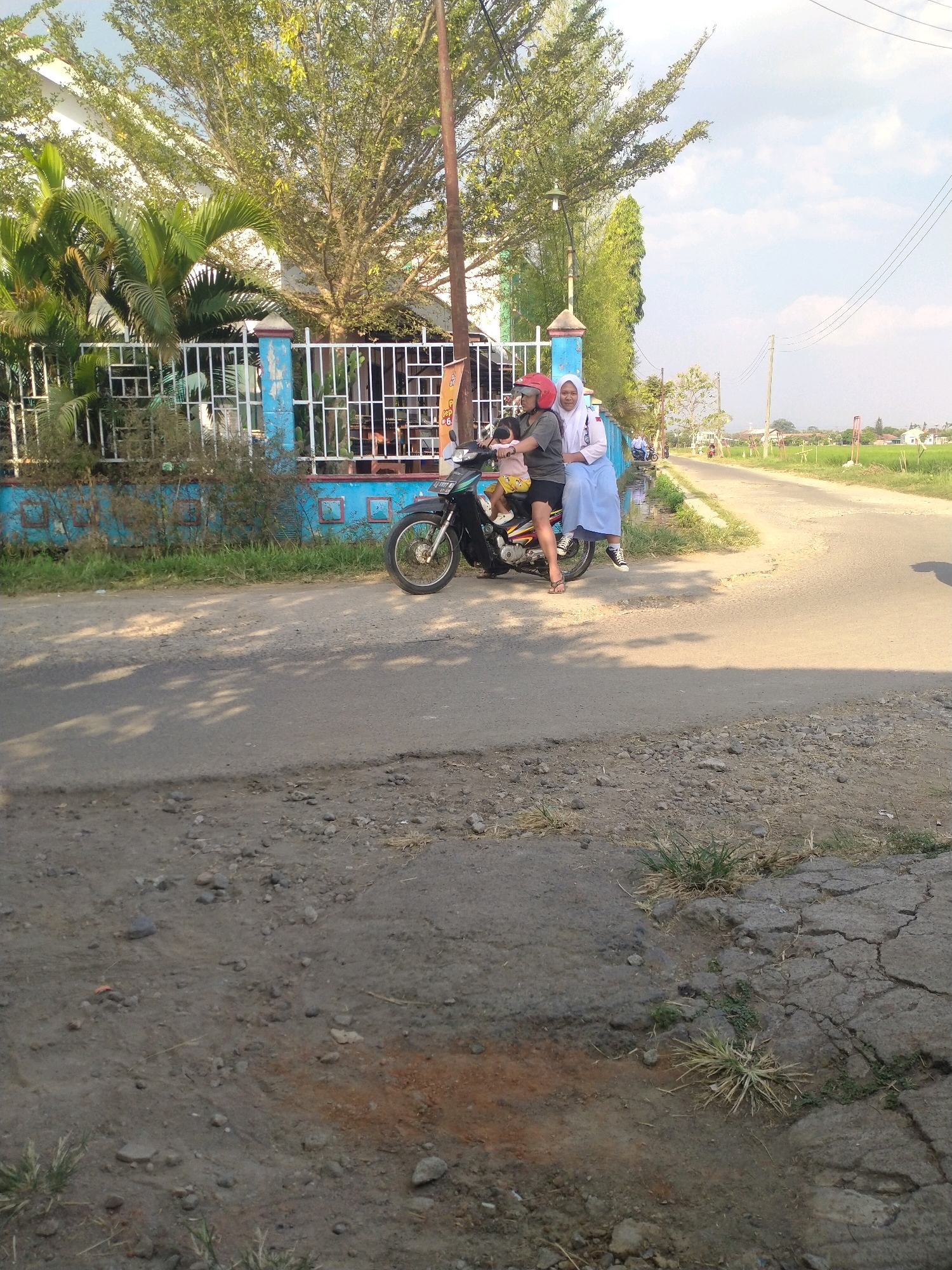 In the city of Begajah, Indonesia, three individuals, two adults and a child, are seen on a motorcycle parked on a dirt road adjacent to a blue and white fence. The adult in the driver's seat wears a red helmet and a dark shirt, while the adult passenger beside them is dressed in a white hijab and a light blue skirt. The child, positioned in front of the adult driver, wears a yellow shirt and is seated on the motorcycle. The surrounding environment features lush green trees, a tall pole with a lamp, and power lines overhead. A paved road stretches into the distance, bordered by green fields and houses, indicating a semi-rural setting. The ground in the foreground is rough, with visible dirt, stones, and patches of dry grass.