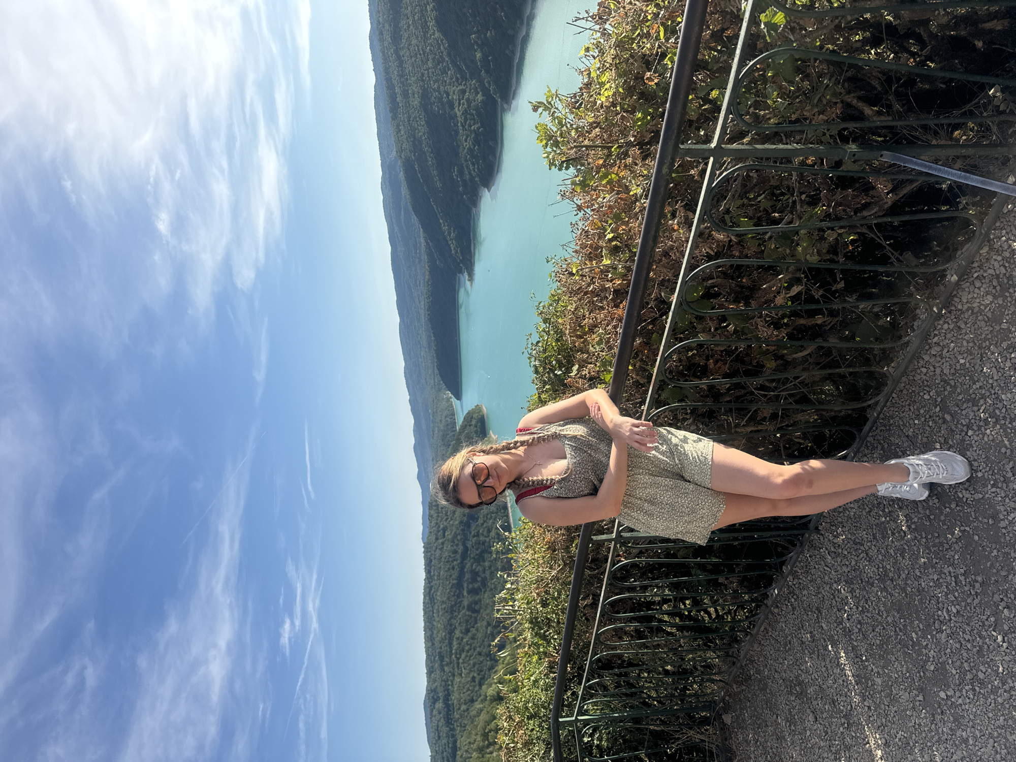 A woman stands on a gravel pathway, leaning against a dark metal railing. She is wearing a floral print romper, sunglasses, and white sneakers. Behind her, a large turquoise lake is surrounded by lush green hills. The sky above is a clear blue with scattered white clouds. The location is the Lac de Vouglans, a reservoir located in the Jura department of France, near the commune of Moirans-en-Montagne.
