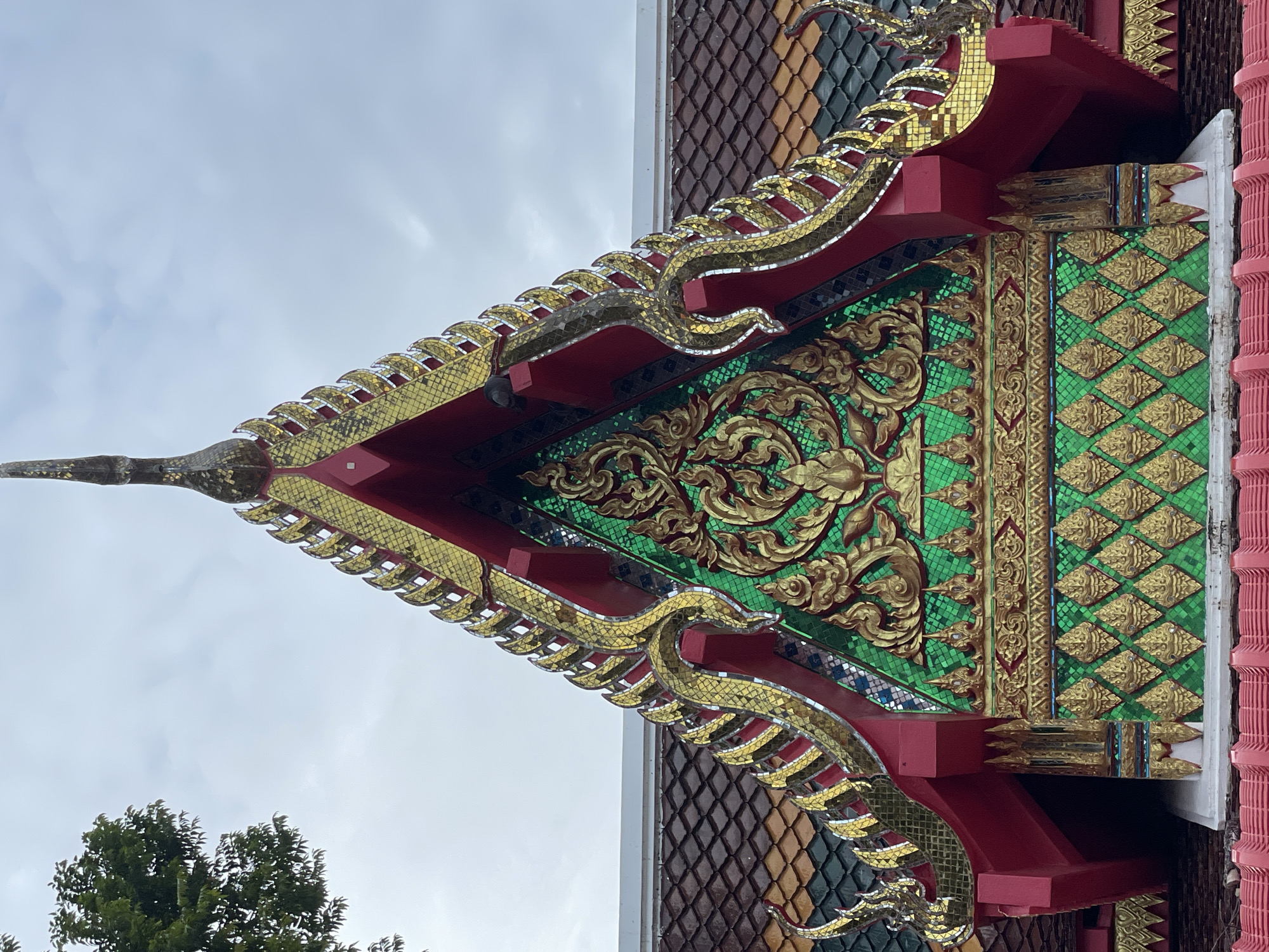 The image shows a close-up view of the ornate roof of a Thai temple in Pattaya City, Thailand. The roof is decorated with vibrant colors and intricate gold leaf detailing, featuring traditional Thai architectural elements such as tiered eaves and decorative finials. The central gable is adorned with a large, gilded floral motif set against a backdrop of emerald green mosaic tiles. The surrounding roof tiles are a mix of dark brown and green, adding to the rich texture of the structure. A portion of a lush green tree is visible in the upper left corner, contrasting with the man-made beauty of the temple. The sky is overcast with soft, diffused light. No people are visible in the frame.
