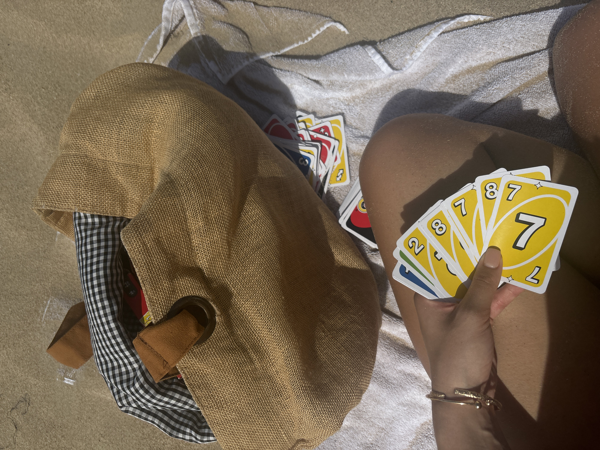 A person's legs are visible, with a beach towel and a straw bag on a sandy surface. The person is holding a hand of Uno cards, and more Uno cards are scattered near the bag. The straw bag has a checkered fabric lining and brown leather handles. The scene appears to be outdoors on a sunny day, likely at a beach, but the specific location in San Juan, United States of America, cannot be determined from the visual information provided.