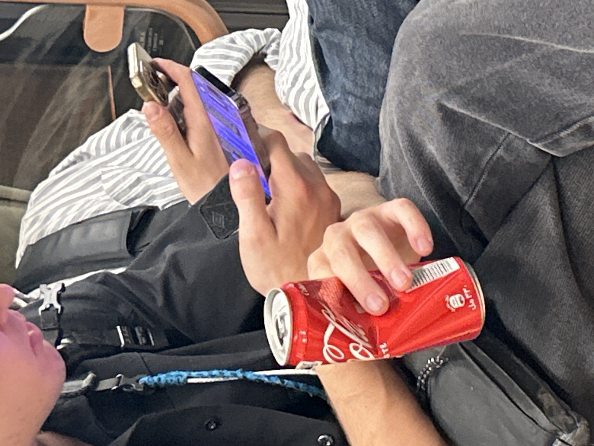 A mid-level shot displays two individuals seated close together, one holding a crushed Coca-Cola can and the other holding two cell phones. The person holding the can has a black shirt with silver buckles, a blue patterned strap, and a silver bracelet. The can is red and white, with the label "Coca-Cola". The other person is using one phone, with a gold frame and what appears to be a purple screen, while holding another phone with a gold back. The individual also wears a striped shirt, a black jacket, and grey jeans. The background is blurred, suggesting an interior setting. The location in the city is unknown, unknown.