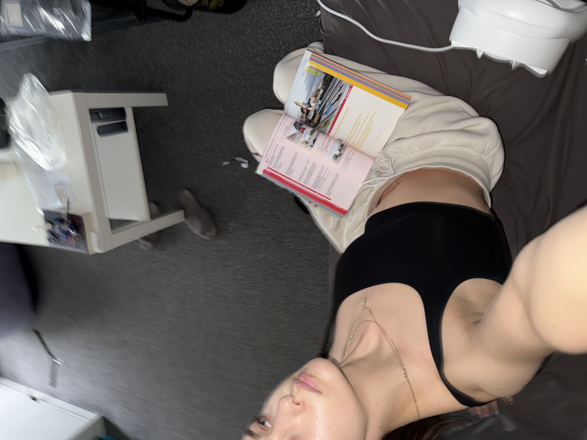 A selfie taken from a high angle shows a person reclining on a bed with a brown sheet. The person is wearing a black top and light-colored sweatpants. They are holding two travel guides open on their lap. A white, cylindrical object with a cord attached is visible on the bed. A white side table is on the left, with some objects partially obscured by a plastic bag on top. The floor is gray carpet. Given the travel guides, the location might be related to travel planning or tourism, but there is no definitive element to pinpoint Sofia, Bulgaria, or any specific city.