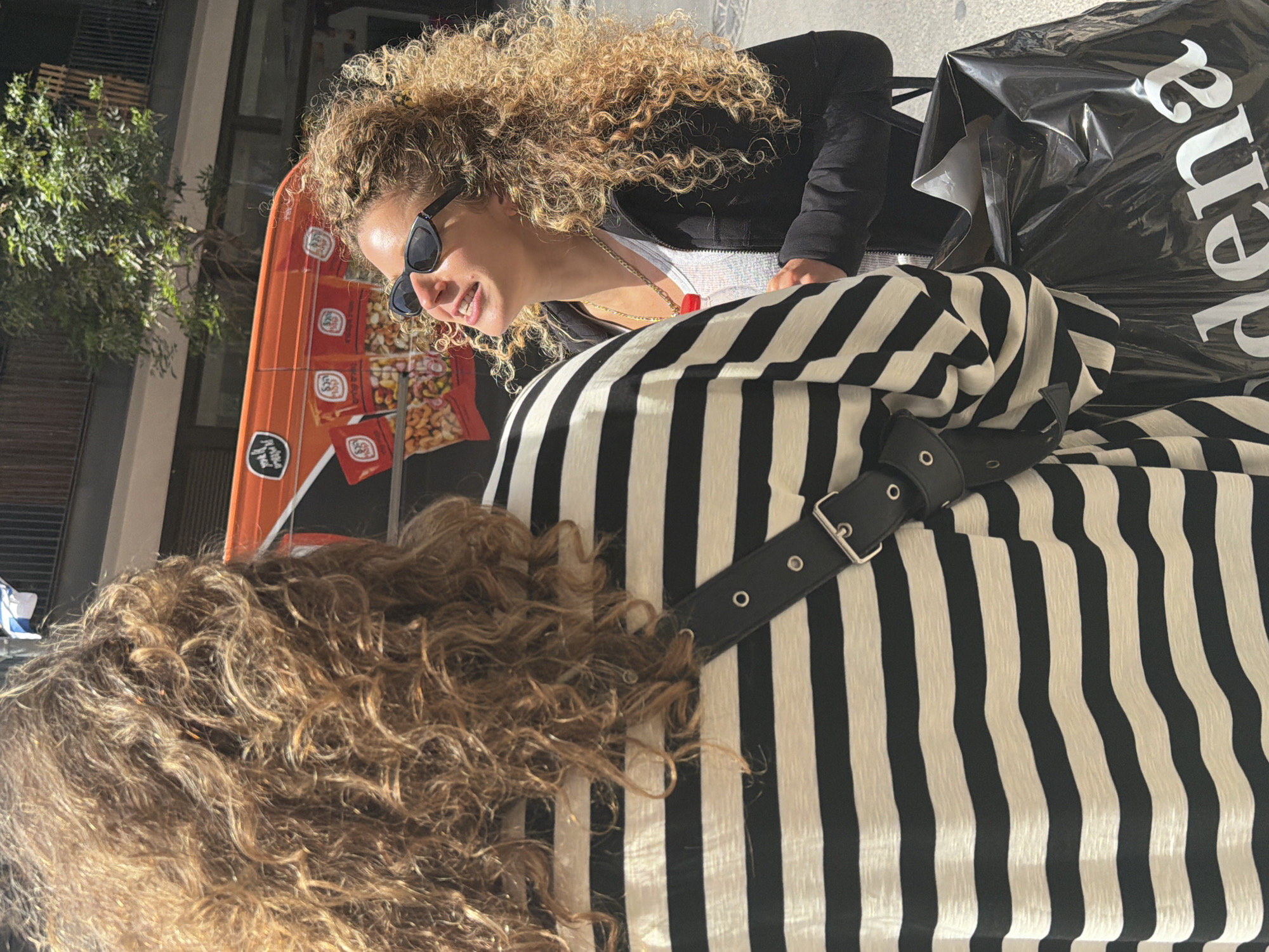 The image depicts two individuals in an outdoor setting in Tel Aviv, Israel. One person, with curly blonde hair and wearing sunglasses and a black jacket, is positioned facing to the left and smiling. A necklace with a red pendant is visible. The second person, primarily seen from the back, has curly brown hair and is wearing a black and white striped top with a buckled black belt. An orange advertising stand with images of nuts can be seen in the background. A black plastic shopping bag with white lettering is also visible, indicating the word "Rena". A tree is visible in the background. The setting appears to be a street or public space in Tel Aviv.
