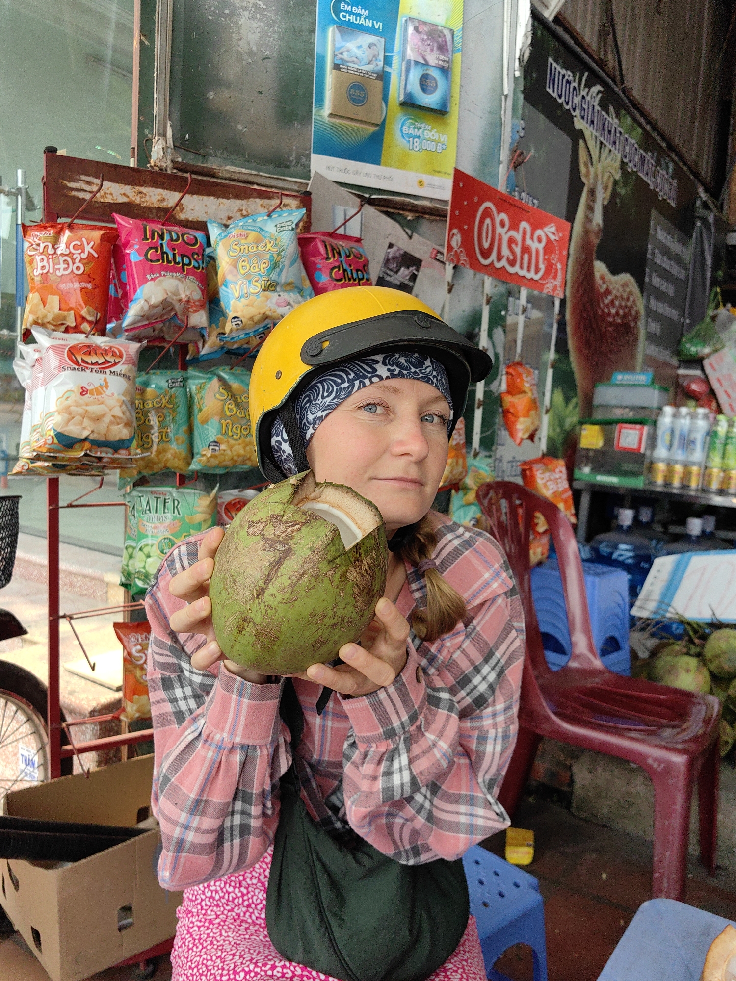 The image shows a person, presumably a tourist, holding an opened coconut in their hands. They are wearing a yellow helmet with a black visor, along with a patterned headband beneath. The person is dressed in a pink plaid shirt over a pink floral dress, and a green bag is slung across their shoulder. The background reveals a street-side shop in Cao Xanh Ward, Vietnam. The shop shelves are stocked with various snacks including brands like "Snack Bi Đỏ," "Indo Chips," "Oishi," and "Snack Bắp Vị Sữa". There are also drinks and other items like coconuts visible. There are promotional posters and signs on display, including an ad for "555" cigarettes. There are red plastic chairs. A bicycle wheel is visible at the left edge of the frame.