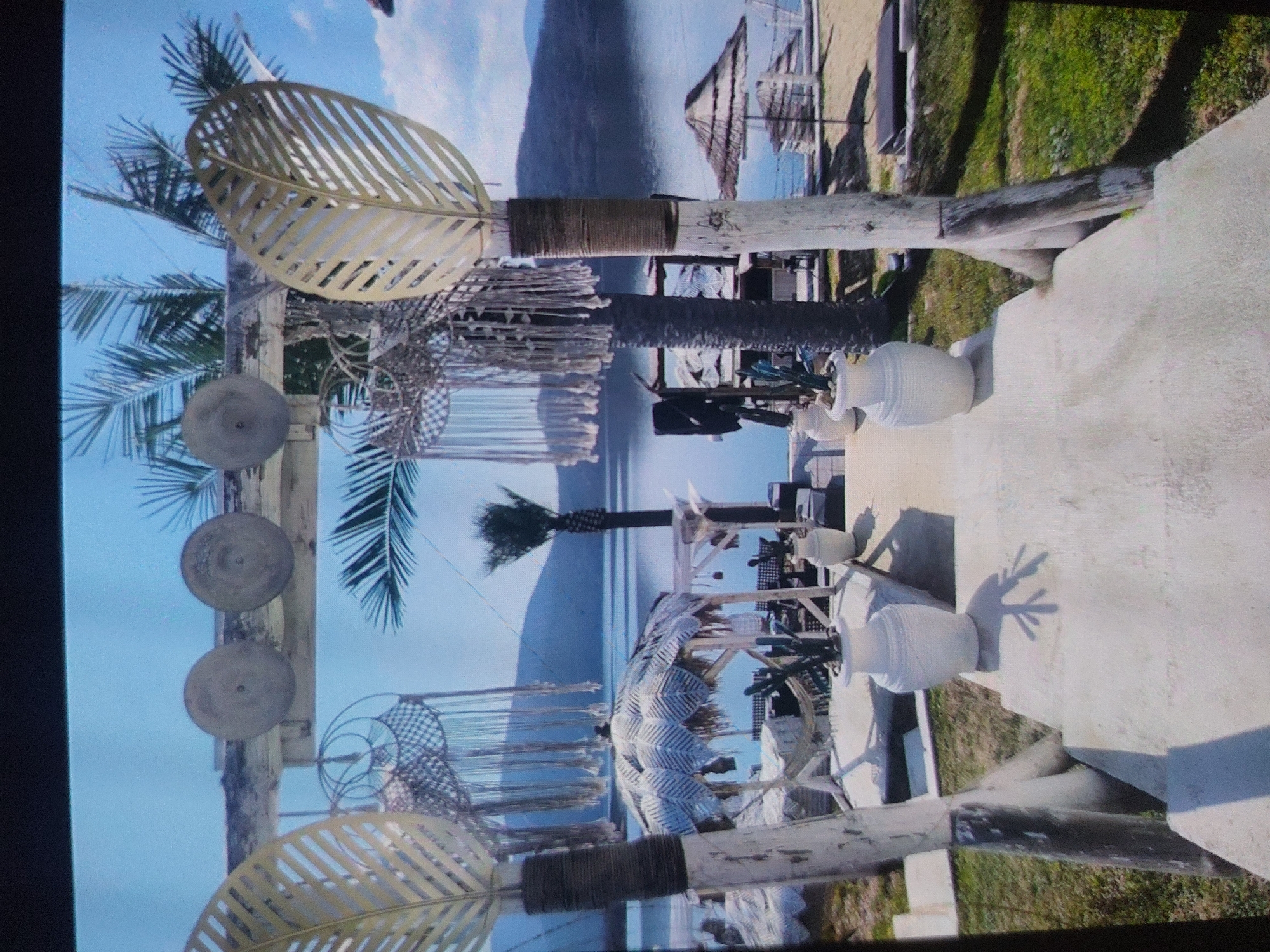 This media file captures a bright outdoor scene, likely a resort or beachside establishment, positioned next to a large body of water under a clear blue sky. The image appears to be rotated 90 degrees counter-clockwise.
The setting suggests a warm, sunny location, consistent with Tsarevo, Bulgaria. In the foreground, on what appears to be a white concrete or paved area, are several white, urn-shaped decorative vases. Long shadows are cast across this surface, indicating bright sunlight, likely during the late morning or early afternoon. To the left and right, rustic wooden posts wrapped with dark rope stand vertically, supporting horizontal beams. These posts are adorned with large, golden or bronze-colored metal cut-outs shaped like palm leaves, and also feature round, flat, grey disks and intricate macrame or woven hanging decorations.
In the mid-ground, multiple palm trees are visible, varying in height, adding to the tropical ambiance. Behind the foreground structures, there are more white, possibly canvas or straw, woven sunshades or shelter structures, indicative of seating or relaxation areas.
The background prominently features a vast expanse of calm, blue water, likely the sea or a large lake. Across the water, distant land or mountains are faintly visible under the clear horizon. No people are visible in the image, suggesting a quiet moment at the location. There is no visible text. The overall atmosphere conveyed is one of relaxation and holiday.