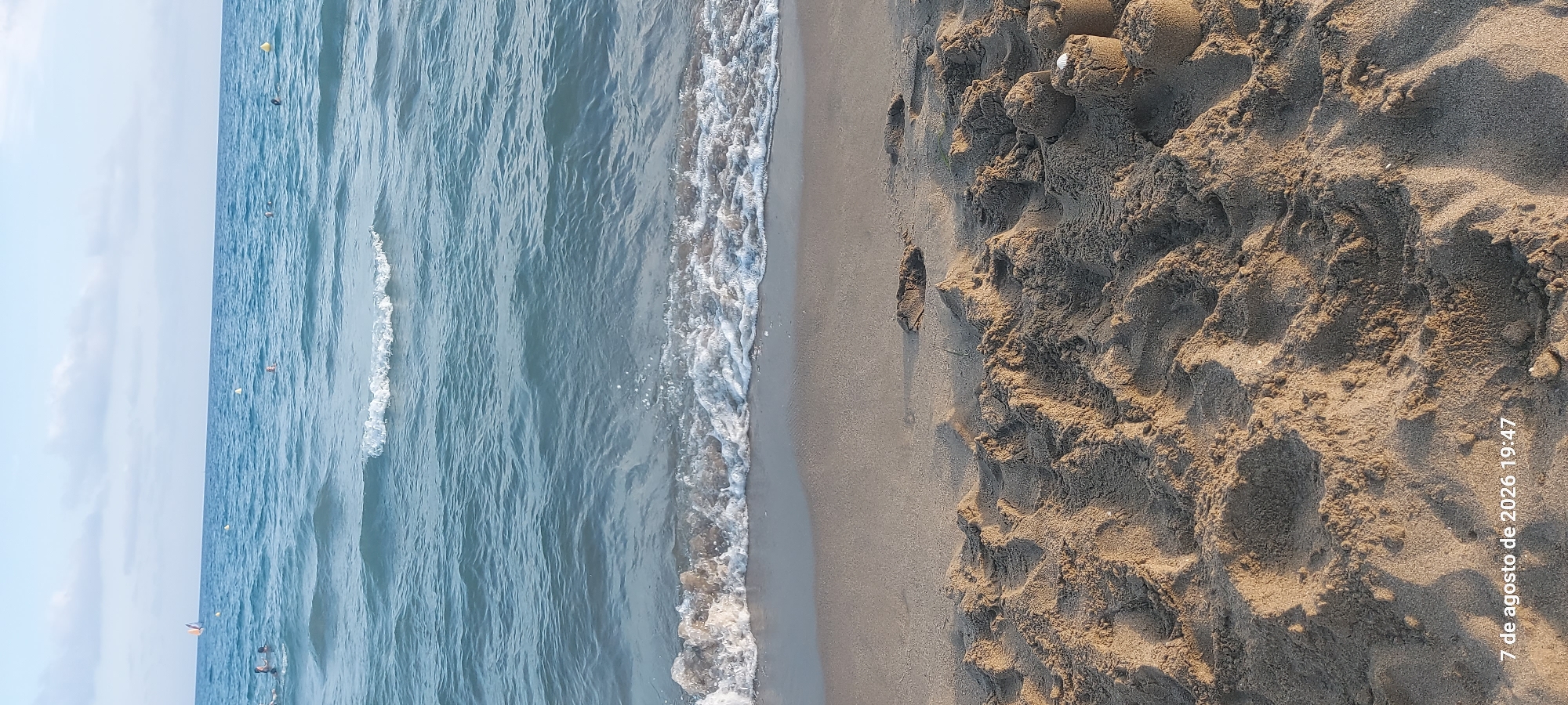 The image shows a beach scene in Castelldefels, Spain, with the Mediterranean Sea in the background. The foreground is dominated by sand, featuring sandcastles and footprints. The sea is calm with gentle waves lapping at the shore, and several people are visible swimming or wading in the water. The sky is partly cloudy, suggesting it is a pleasant day. A date and time stamp, "7 de agosto de 2026 19:47", is visible in the lower left corner, indicating the image was taken in the late afternoon. The overall impression is one of a relaxed summer day at the beach.