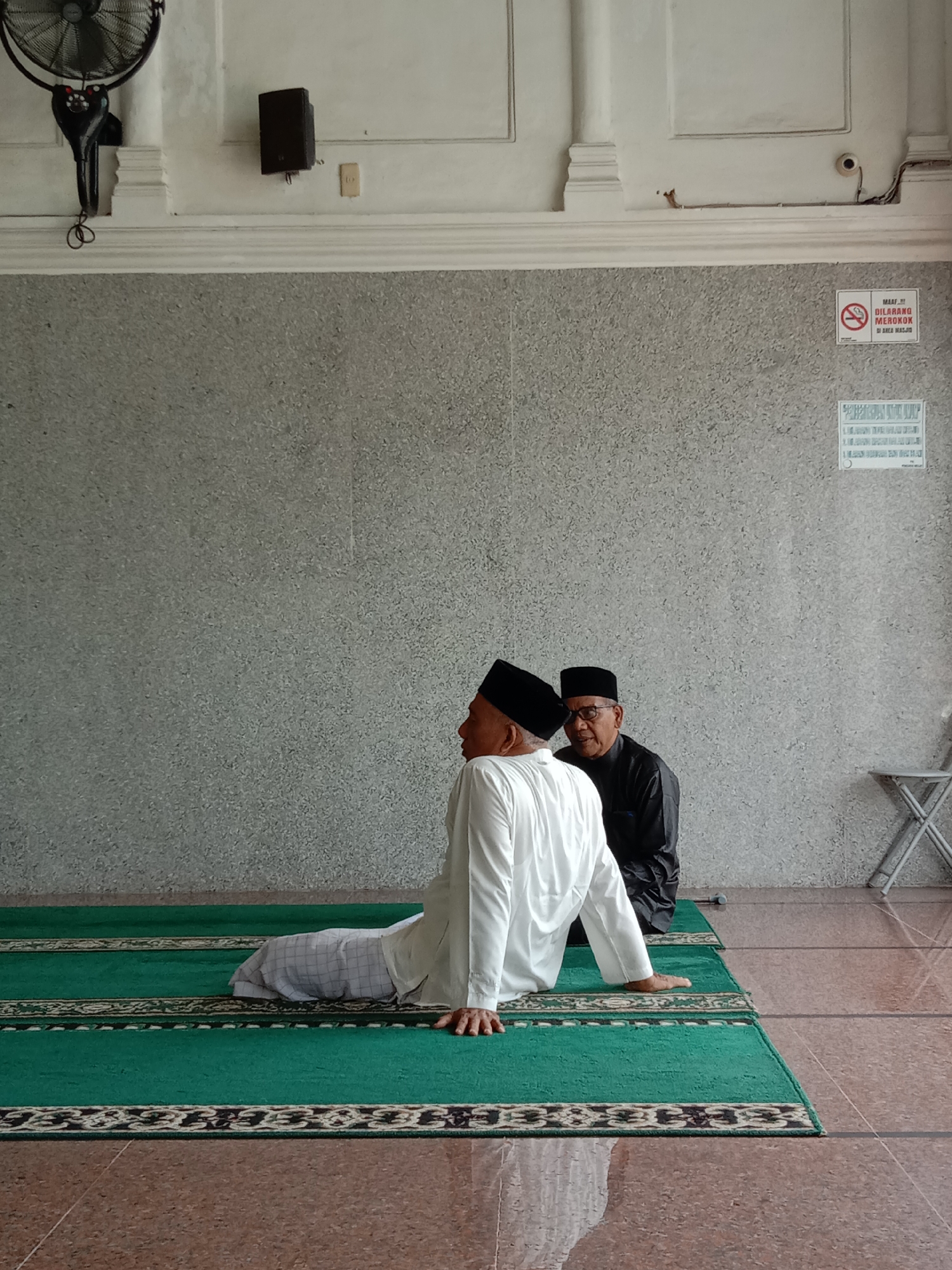 Two men are sitting on a green prayer rug laid on a reddish-brown tiled floor inside a building. The man in the foreground is wearing a white outfit and a black cap. He is leaning back on his hands. The man in the background is wearing a dark outfit and glasses, also with a black cap. They are in Lhokseumawe, Indonesia. The wall behind them is gray. A fan is mounted in the upper-left corner of the wall. There is a speaker, a "No Smoking" sign, and a calendar hanging on the wall. There is a small metal folding chair near the wall in the right background.