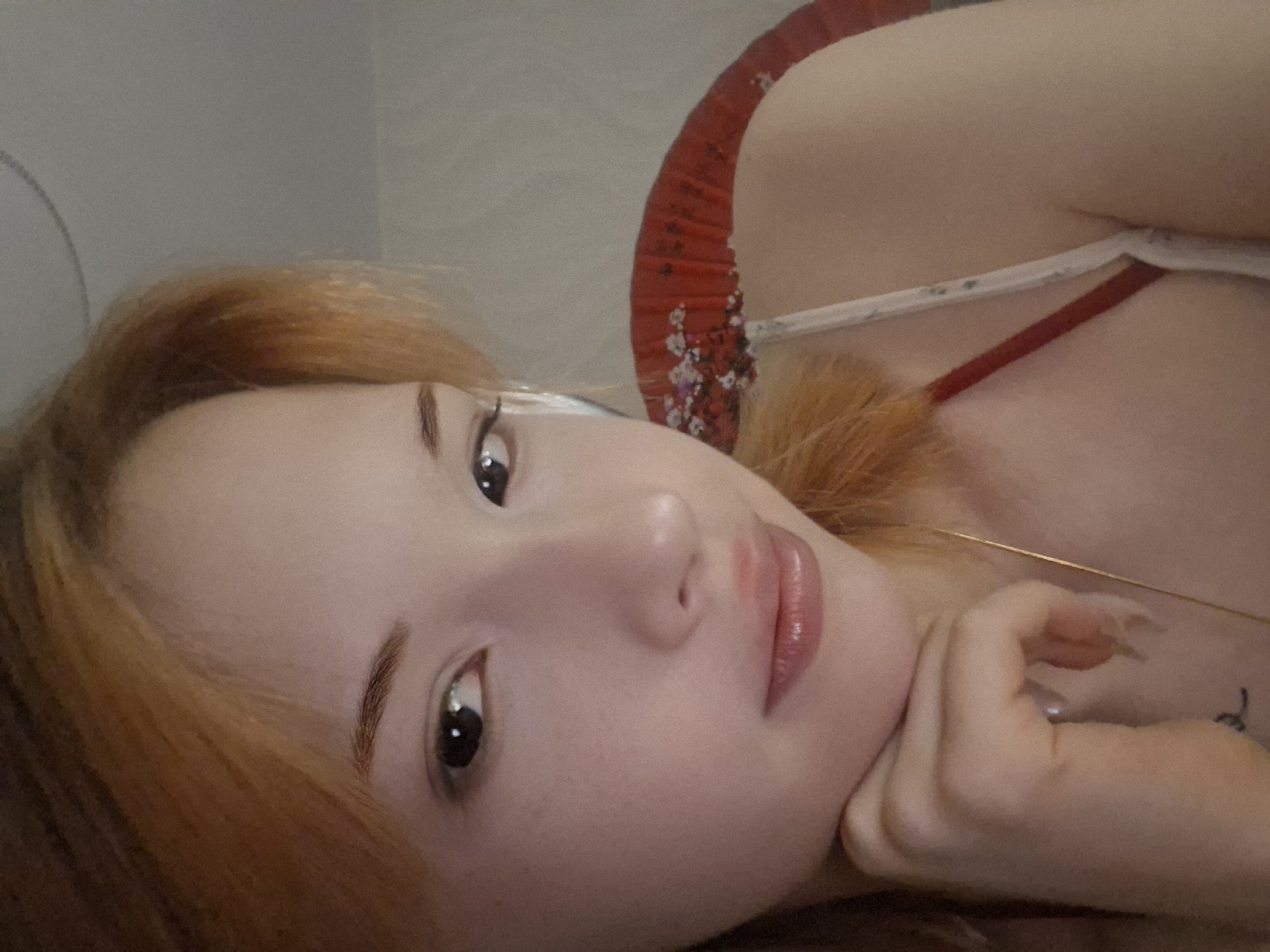 A close-up, slightly tilted portrait captured in Chaikovsky, Russia, features a young woman with fair skin and long, reddish-orange hair. She is looking directly at the camera with dark eyes and a neutral to slightly contemplative expression, her lips appearing slightly parted and glossy. Her right hand is gently resting under her chin, revealing long, light-colored fingernails.
Behind her head and shoulders, a vibrant red, fan-like object with white or silver decorative accents is visible, possibly a traditional Asian fan or a decorative element of clothing. A thin, golden chain or necklace can be seen draped across her chest. A white bra strap is visible over her right shoulder. A small, dark tattoo is partially visible on her right forearm or shoulder area. The background is a plain, light gray or white, suggesting an indoor setting, possibly a wall or ceiling, indicating she might be lying down. The overall lighting is soft and even, typical of indoor conditions. There is no visible text in the image.