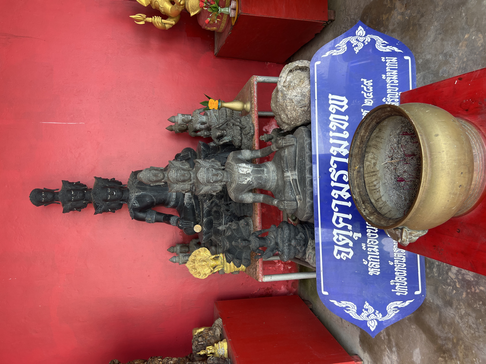 In Karon, Thailand, a religious shrine is depicted against a vibrant red wall. The shrine features several stone Buddha statues of varying sizes, arranged on a tiered shelf. The largest statue in the center has multiple faces. Smaller statues surround it, some golden, some black. A brass incense burner filled with ash and remnants of incense sticks sits in the foreground on a red surface. A blue sign with white Thai script is placed in front of the burner, detailing religious information. The floor is concrete.