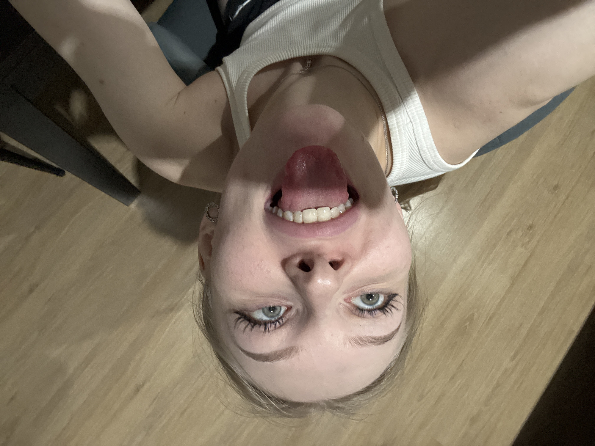 A close-up, upside-down selfie taken indoors shows a fair-skinned person with light blonde hair, blue eyes, and visible teeth. They are wearing a white ribbed tank top and silver hoop earrings, sticking their tongue out towards the camera. The background consists of a wooden floor and a dark object, likely a table or chair leg, is partially visible in the corner. The location in the city of Perm, Russia is unknown.