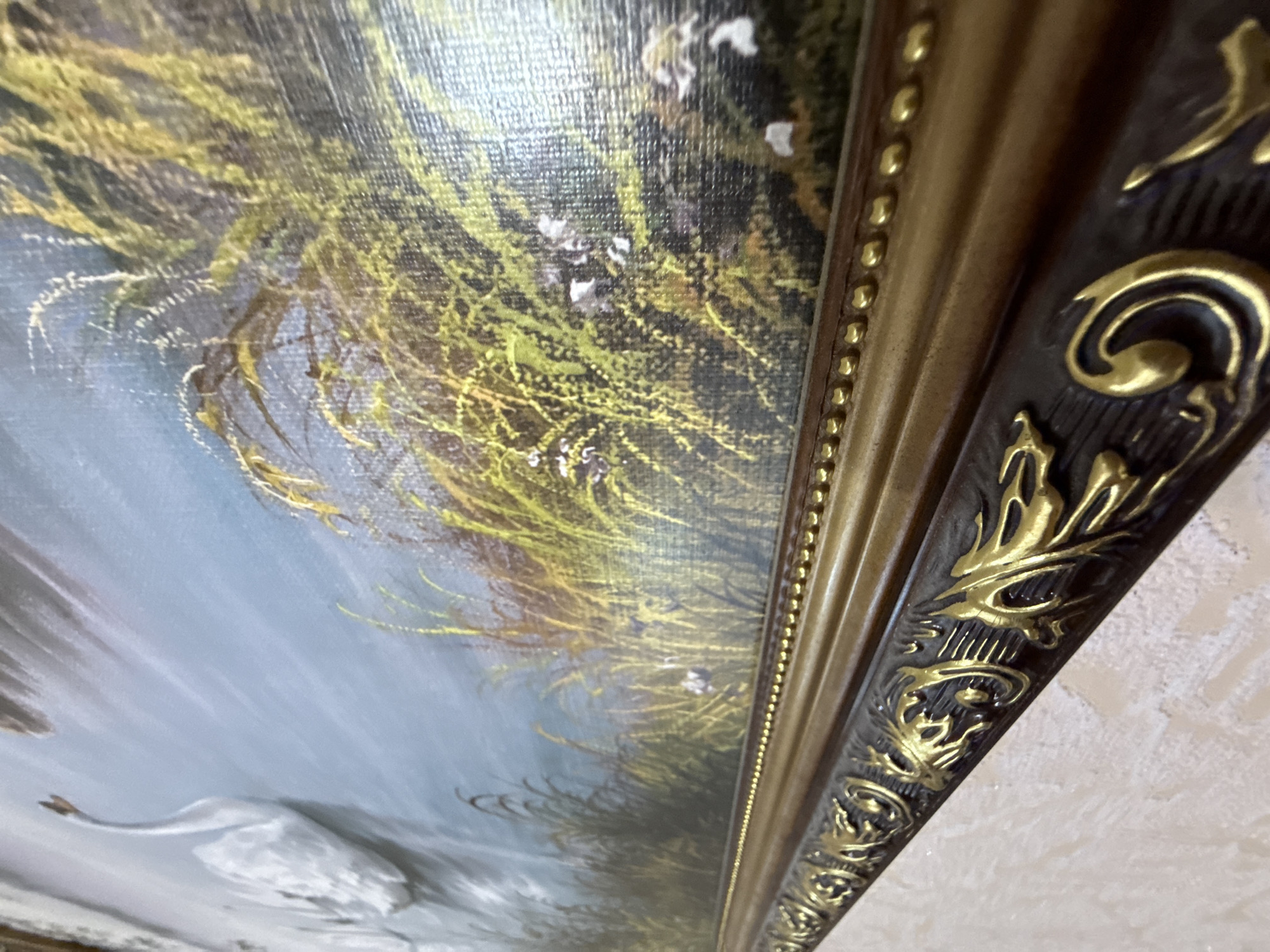 This is a close-up, angled shot of a framed painting. The painting itself appears to be a landscape scene, with a body of water or a river dominating the foreground, rendered in muted blues and grays. The water shows some white reflections, suggesting movement or light. On the left side of the painting, a white swan is partially visible, with its neck elegantly curved. The background of the painting is filled with dense, reedy grasses in shades of yellow and green, with some small white flowers interspersed. The painting is housed in an ornate frame. The frame has a dark brown base with intricate gold detailing. The gold ornamentation includes swirling patterns and leaf-like motifs, and a beaded border runs along the inner edge of the frame. The texture of the canvas is visible, indicating it is likely an oil or acrylic painting. The lighting in the image suggests it might be daytime, possibly with soft light, given the way light reflects on the water and the frame. There is no visible text in the image. The overall impression is of a classical or decorative artwork, focusing on a serene natural scene.