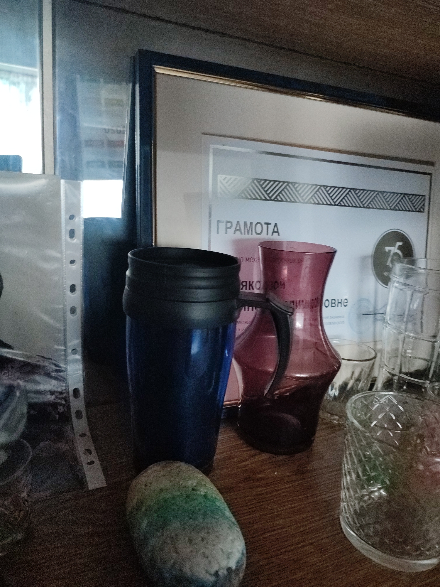 This is a close-up, eye-level, indoor photograph of a wooden shelf. On the shelf, from left to right, are a transparent plastic document holder, a dark blue travel mug, a decorative stone with green and yellow marbling, a pink glass pitcher, and several stacked clear glass tumblers. Behind the objects, a framed document with text in Cyrillic script is leaning against the wall. The document appears to be a certificate or award, with decorative geometric patterns. The environment suggests a home or office setting. No specific location in Narowlya, Belarus, is identifiable from the image.