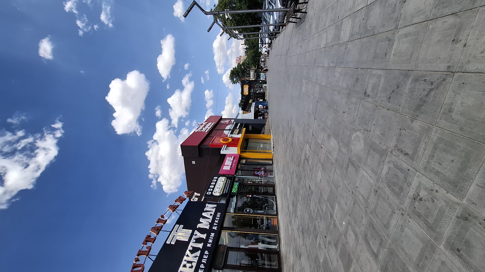 A wide-angle, eye-level, vertical shot shows a pedestrian walkway lined with shops on the left and parked cars and trees on the right, under a bright blue sky with scattered white clouds. The pavement in the foreground is composed of large, square grey tiles, some with decorative borders.
On the left, a row of storefronts displays various signage. The leftmost sign reads "SULTAN" in large, red, serif letters above "EKTY MAN" in white, serif letters with a "T" logo. Below "EKTY MAN" in smaller text is "ЕРЛЕР КИІМ ДҮКЕНІ". Next to it, a store called "REMAX" is visible, with smaller signs indicating "AUDIO & ACCESSORIES" and "REPAIR SERVICE CENTER". Another store has "ILIKЕ" in pink and yellow lettering, with "burger & grill" below it. Further down, a red storefront with white lettering says "ALI", and next to it, "ZRZA".
In the distance, more shops are visible, including one with "MOOZ" in bright letters, and further on, a sign for "AQUA ROSSA". People are seen walking on the sidewalk and entering and exiting the shops. On the right side of the walkway, several cars are parked in a row. The background features a line of trees, indicating an outdoor urban environment. The lighting suggests it is daytime, likely mid-morning or afternoon, with clear weather. The overall scene depicts a commercial area in a city. The specific location is a shopping street in Shymkent, Kazakhstan.