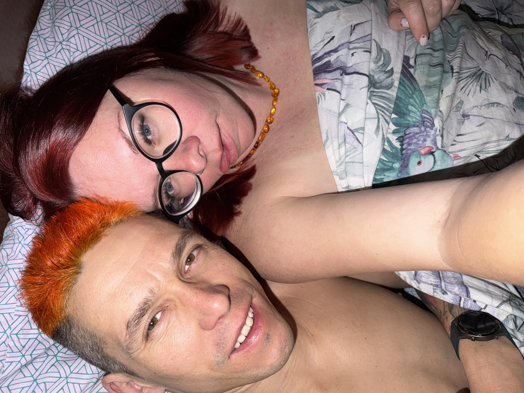 A selfie-style image shows two people lying in bed. The person in the foreground has short, bright orange hair and is smiling, exposing their teeth. The person behind them has reddish-brown hair, is wearing glasses, and a yellow beaded necklace. They are partially covered by a blanket or sheet with a white tropical bird and foliage print. The bed linens underneath have a geometric pattern in white and green. The image was likely taken indoors, possibly in a residential setting in Chaikovsky, Russia, given the prompt's specification. A black smartwatch is visible on one of the persons.