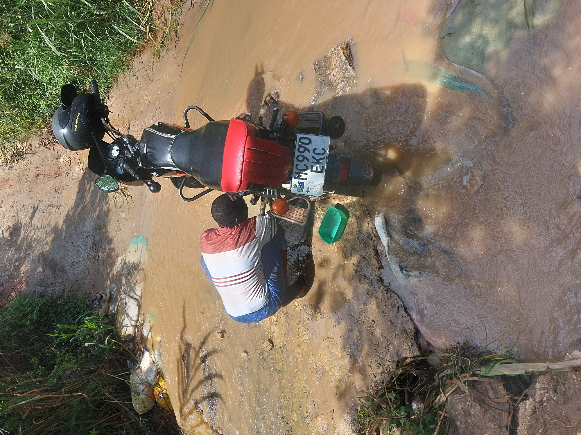 The image captures a scene in what appears to be a rural or semi-rural area, potentially on the outskirts of Dar es-Salaam, Tanzania. A person is kneeling near a pool of muddy water next to a motorcycle. The person is wearing a white t-shirt with red stripes and blue jeans. The motorcycle, with a black and red seat and black helmet, is partially submerged in the water. The license plate reads "MC990 EKC". A green bucket and other indiscernible objects are visible near the person and the motorcycle. The surrounding environment includes dirt, grass, and debris, suggesting an area prone to flooding or heavy rainfall.