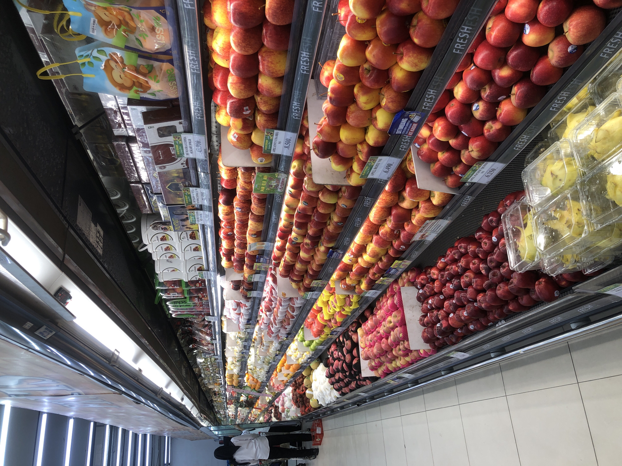 The image depicts the interior of a brightly lit supermarket aisle in Langsa, Indonesia, with a focus on the produce section. Rows of fresh fruits, primarily apples, are meticulously arranged on tiered shelves. The apples vary in color, from deep red to yellow and green, and are presented in bulk as well as pre-packaged containers. Some shelves have price labels. In the background, other produce like grapes and possibly pears are visible, as are packaged goods and promotional displays. Two individuals, likely shoppers, are partially visible at the far end of the aisle, one wearing a white shirt and dark pants, and another in a dark hijab and dark clothing. The overall environment suggests a well-stocked and organized retail space.