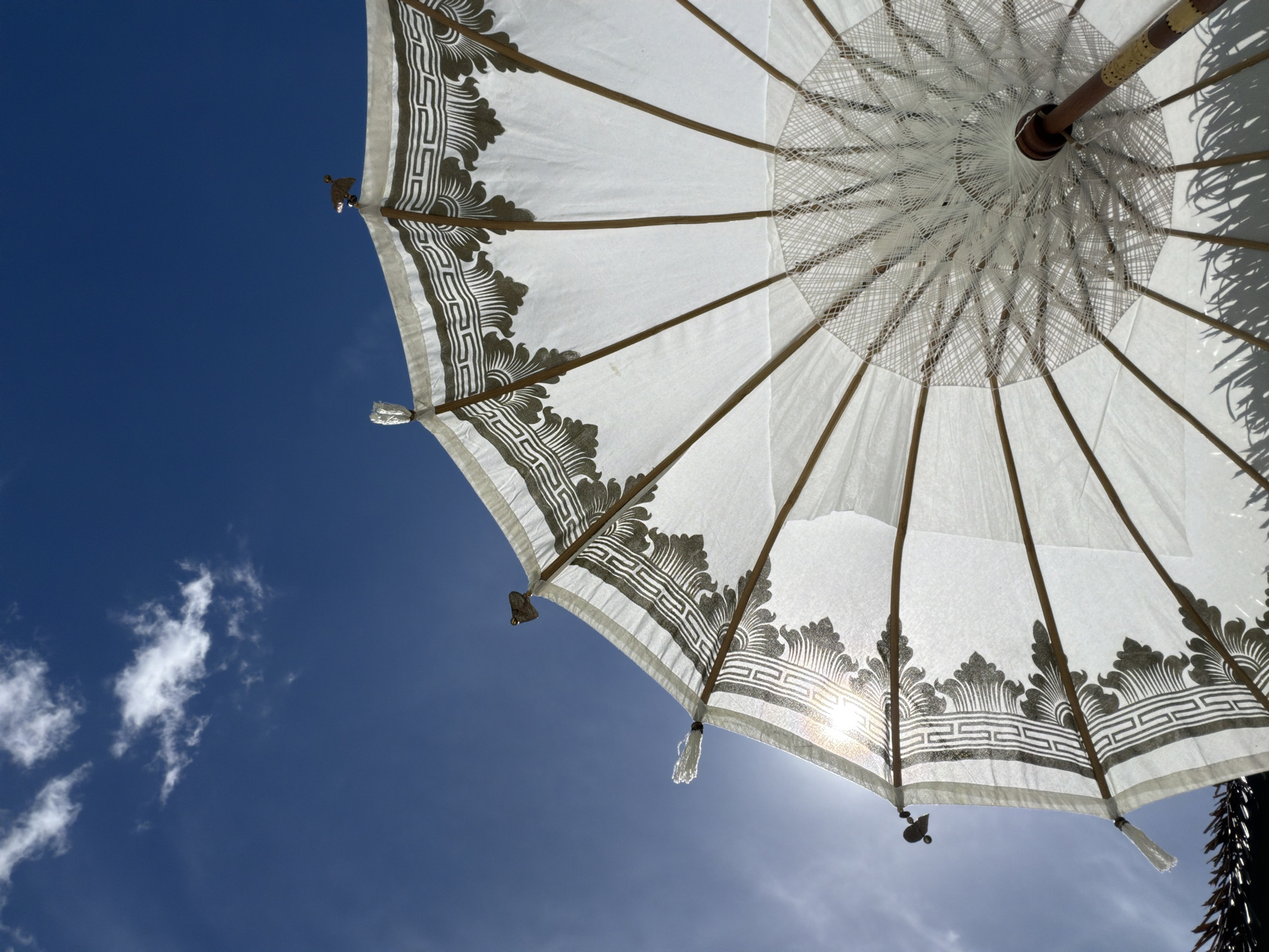 An upward-angle shot captures the underside of a white, ornate umbrella against a deep blue sky with sparse white clouds. The umbrella is constructed with thin wooden ribs and a delicate white fabric canopy, adorned with a dark, repeating geometric and floral pattern along its edge. The sun shines through the translucent fabric, creating a bright glare and casting dappled shadows of what appear to be palm fronds at the bottom of the frame. Small, decorative metal bird figures are perched on the tips of some of the umbrella's ribs. The scene suggests a bright, sunny day in a tropical location. The image was taken in Baan Bo Phut, Thailand.