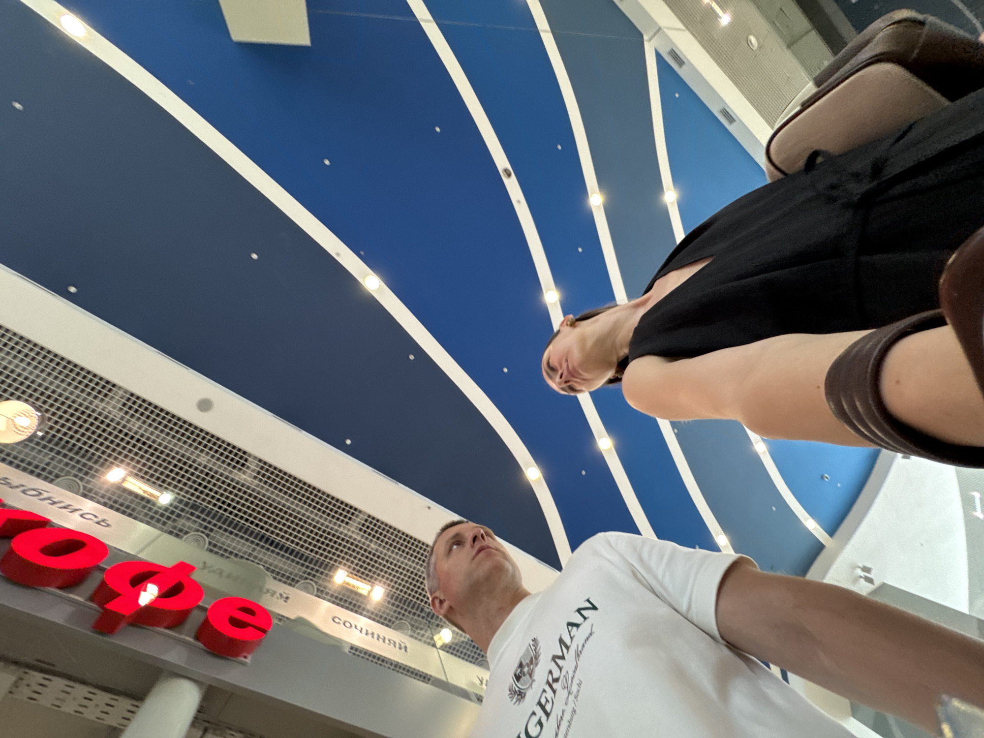 A low-angle, upward-looking photograph captures a man and a woman in an indoor public space in Sochi, Russia. The man, positioned on the left, has short, light-colored hair and wears a white long-sleeved t-shirt with "GERMAN" and "St. Petersburg / Sochi" printed on it. He looks slightly upwards and to his right with a neutral expression. The woman, partially obscured by her own arm and hand in the foreground, is positioned on the right. She has dark hair, wears a black V-neck top, and looks upwards and to her left with a serene expression, a small light-colored earring visible on her left ear. She carries a light brown shoulder bag on her right side. Both individuals appear to be observing the high ceiling.
The ceiling features a design with alternating bands of dark and medium blue, separated by white curvilinear stripes. Numerous small, circular recessed lights are embedded within these colored sections, emitting a warm glow. Above the man, a red, illuminated Cyrillic sign with large letters spelling "еде" (ede) is visible against a reflective grid ceiling. Below this grid, reflections show Cyrillic text including "СОЧИНЯЙ" (Sochinyay) and "СОЧИ" (Sochi). The environment suggests a modern commercial or public building, such as a shopping mall or large hall.