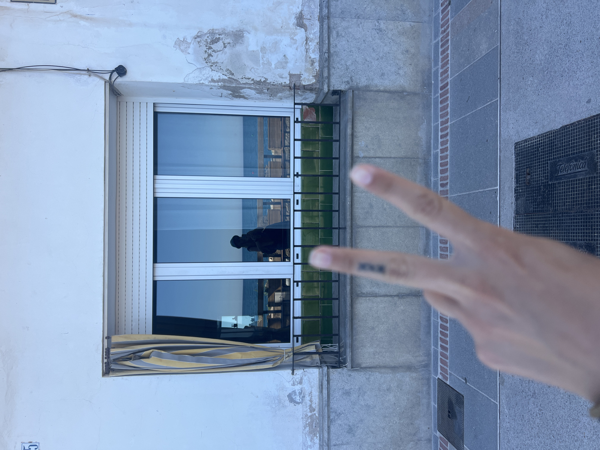 The image shows a building exterior in Sant Pol de Mar, Spain. The wall is partially white with weathered patches, and partially gray. A window with a white frame and closed white roll-down blinds is visible. A striped awning is partially rolled up below the window. A black metal railing sits outside the window. A person's hand is in the foreground, displaying a peace sign. The reflection in the window shows a silhouetted figure inside the building. A gray tiled sidewalk with a small, dark metal utility access cover (Telefonica) is beside the building. The overall setting is outdoors in daylight.