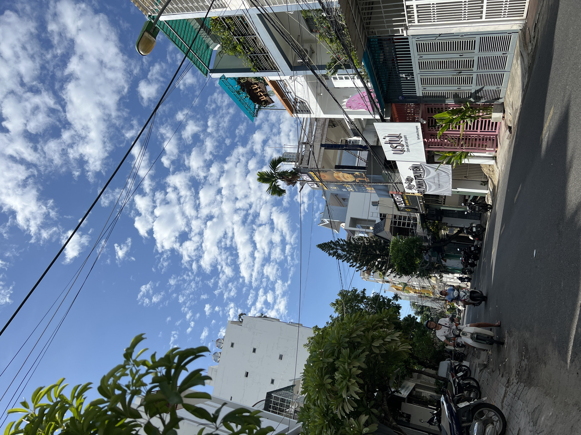 In Nha Trang, Vietnam, on a sunlit day, a street scene unfolds with a clear blue sky dotted with fluffy white clouds. The road is lined with multi-story buildings exhibiting a mix of architectural styles, many featuring balconies and awnings. Several motorcycles are parked along the side of the street, and two individuals are seen riding motorcycles in the middle of the road. One person is on a light-colored scooter in the foreground, while another is on a darker scooter further down the street. Signs in Vietnamese are visible on the buildings, indicating various businesses. Lush green trees and foliage are interspersed among the buildings, adding a touch of nature to the urban environment. Overhead, power lines crisscross the sky.