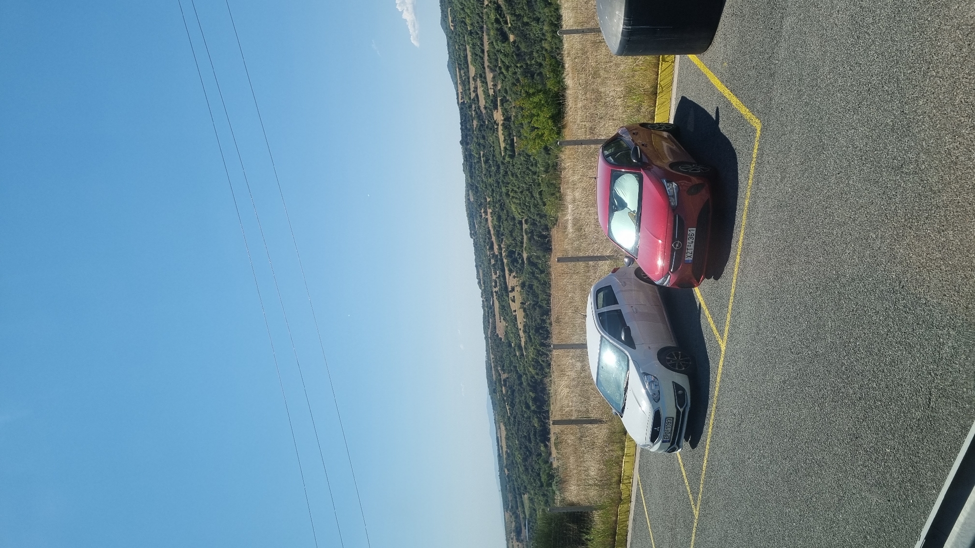 In Strymoniko Municipal Unit, Greece, two cars are parked in a lot. A white hatchback is parked in the left space, and a red hatchback is parked in the adjacent right space. Both cars are facing forward. The parking lot is paved with asphalt and marked with yellow lines. Beyond the parking lot, a fence separates it from a field of dry grass. In the background, a verdant, hilly landscape stretches under a clear blue sky. Power lines are visible against the sky.