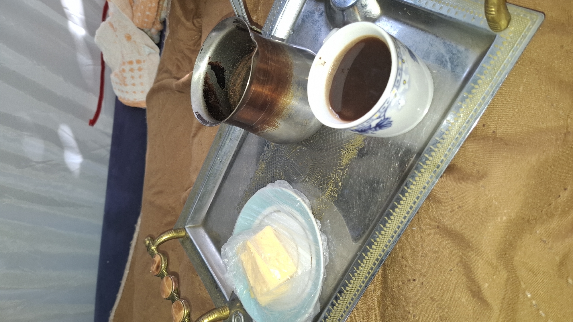 A silver tray with ornate gold trim rests on a brown, textured surface, possibly a blanket or fabric. On the tray, to the left, is a small light blue plate covered with clear plastic wrap, containing two rectangular pieces of yellow cheese. To the right of the cheese is a traditional metal coffee pot, stained with use, with a dark liquid inside. In front of the coffee pot is a white cup with blue floral patterns, filled with dark coffee. The background appears to be a tent or canopy made of white fabric, with some blue material underneath. There is no clear indication of the specific location within Шұбарсу, Kazakhstan.