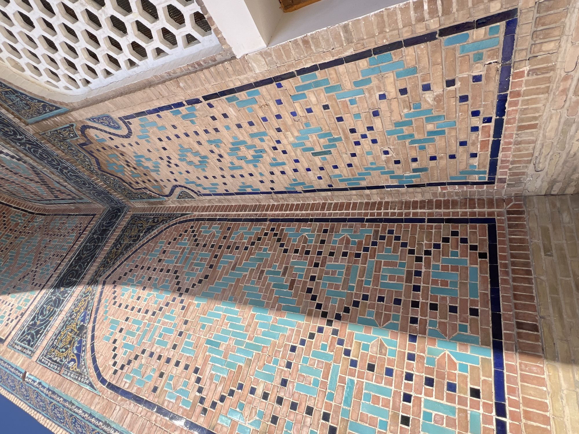 The image shows a close-up of the exterior of a building in Samarkand, Uzbekistan, featuring intricate tilework and brickwork. The wall is adorned with a geometric pattern of small, rectangular tiles in various shades of blue, black, and beige, set against a backdrop of reddish-brown brick. Some sections of the wall are highlighted by the sun, creating a contrast with the shadowed areas. In the upper right portion of the image, a section of a white, honeycomb-patterned lattice is visible, likely part of a window or decorative screen. The overall impression is one of ancient architectural artistry, with the textures of the tiles and bricks clearly discernible.