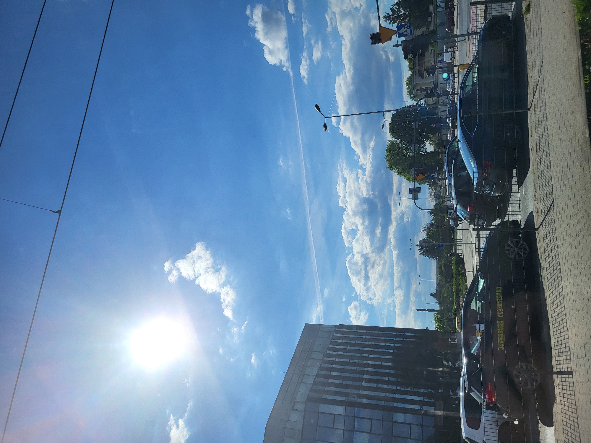 The image shows a bright, sunny day in an urban environment. The sky is predominantly blue, with scattered white clouds. The sun is visible, creating a glare in the lower left corner. A contrail is visible in the sky. On the right side of the image, cars are parked along a sidewalk. Streetlights and traffic signals are also present. A modern building with reflective glass windows is visible at the bottom of the image.