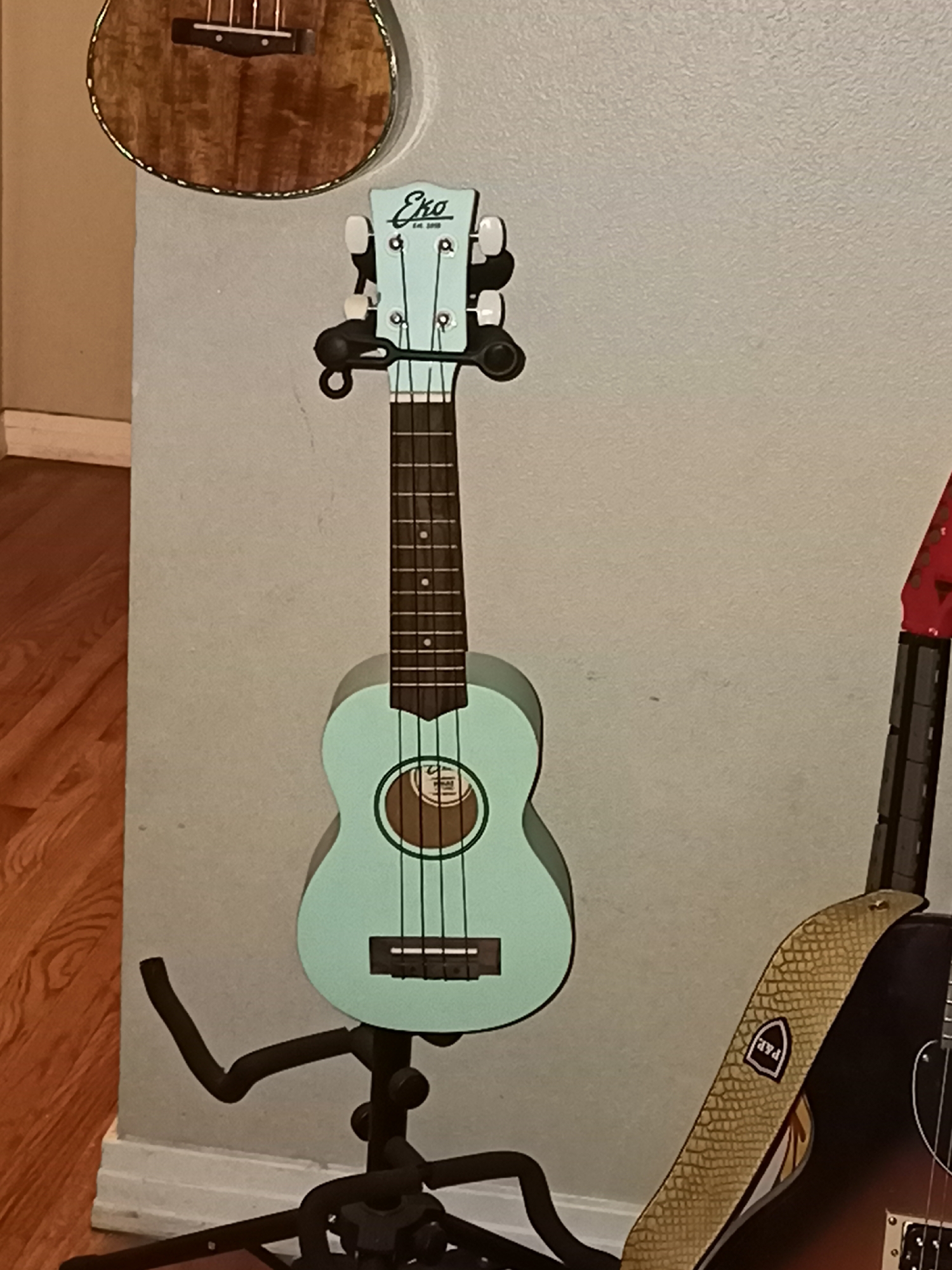 The image shows a light blue ukulele on a black stand, leaning against a pale gray wall. The ukulele has a circular soundhole with a brown inlay. The headstock of the ukulele displays a logo that reads "Eko" and "Est. 2016". To the left of the ukulele, a portion of a wooden ukulele is visible, hanging on a wall. To the right, the neck and body of a sunburst electric guitar with a textured beige strap are visible. The floor is made of polished wooden planks. The scene appears to be indoors, likely a music room or a store display. The location is not specified in the image.