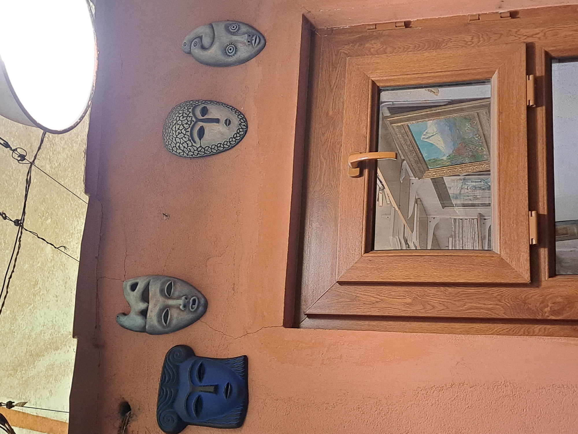 The image shows an exterior wall in Yerevan, Armenia. The wall is painted a muted reddish-orange color and textured. Four face-shaped masks are mounted vertically on the wall. From top to bottom, the masks are gray, gray with a patterned border, gray with an open mouth, and dark blue. To the right of the masks is a light brown wooden window frame, reflecting a room with a painting hanging inside. The window is closed. A bright light fixture is visible on the left side of the frame, and some wires are visible nearby.