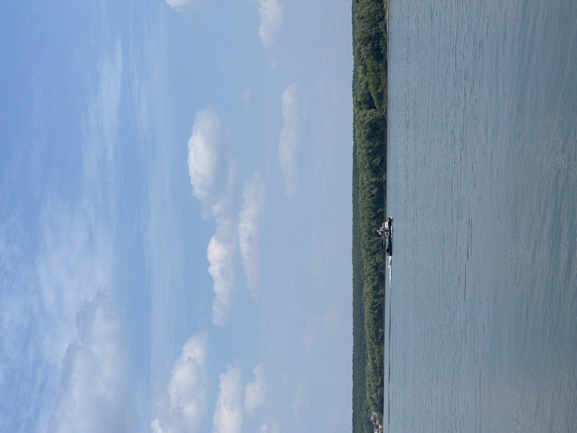 A wide, calm river stretches across the frame under a bright blue sky dotted with fluffy white clouds. A small boat with a dark hull and a lighter superstructure travels from left to right, leaving a faint wake on the water. Along the distant bank, a dense, lush green forest forms a horizontal band. On the far left, a few buildings, possibly houses, are visible nestled amongst the trees, suggesting a human settlement. The overall scene depicts a serene, natural landscape with a touch of human presence.