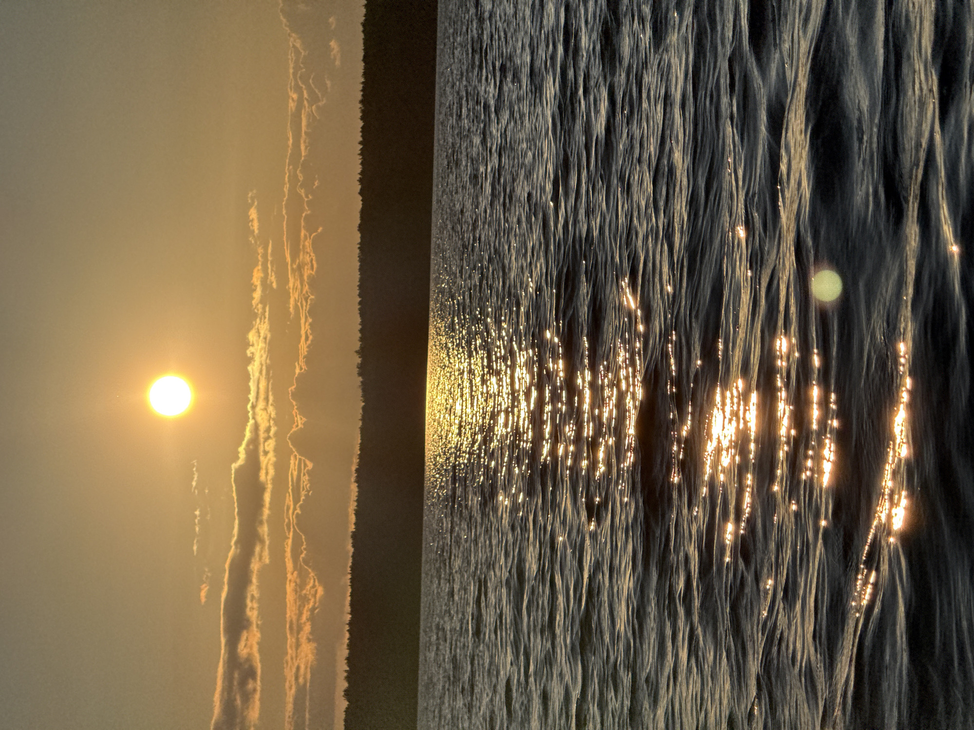 A serene, wide-angle shot captures a tranquil lake at sunset. The sun, a bright, luminous orb, hangs in the upper left quadrant of the sky, casting a warm, golden glow that reflects off the rippling water in a shimmering path. The water's surface is a deep, dark blue, subtly textured by gentle waves. Along the horizon, a dense, dark line of trees forms a silhouette against the luminous sky, suggesting a forested shoreline. The sky itself transitions from a pale, warm yellow near the sun to a softer, dusky hue at the top of the frame, with scattered, elongated clouds tinged with orange and pink. The scene conveys a sense of peaceful solitude and natural beauty, typical of the environment in Gora (Khokhlovskoe S/P), Russia. There are no people or man-made objects visible in the image.