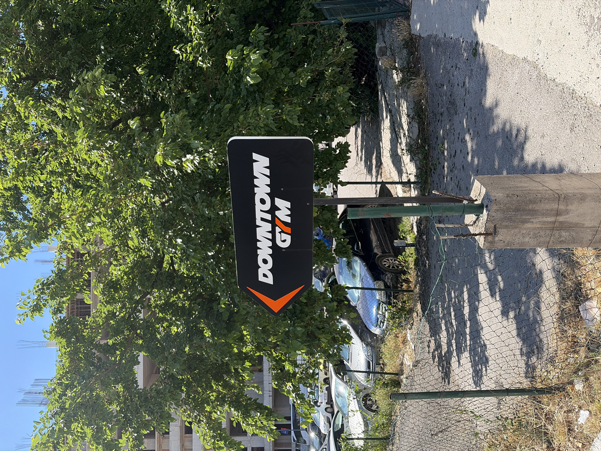 The image depicts a street scene in Budva, Montenegro, on a sunny day. A prominent sign indicates "Downtown Gym," with a black background and orange arrow. The sign is positioned beside a wire fence, adjacent to a parking area filled with cars. A large green tree partially obscures a building under construction with exposed rebar. The area is overgrown with weeds and dry grass. A concrete sidewalk runs along the fence line, casting shadows from the tree overhead.