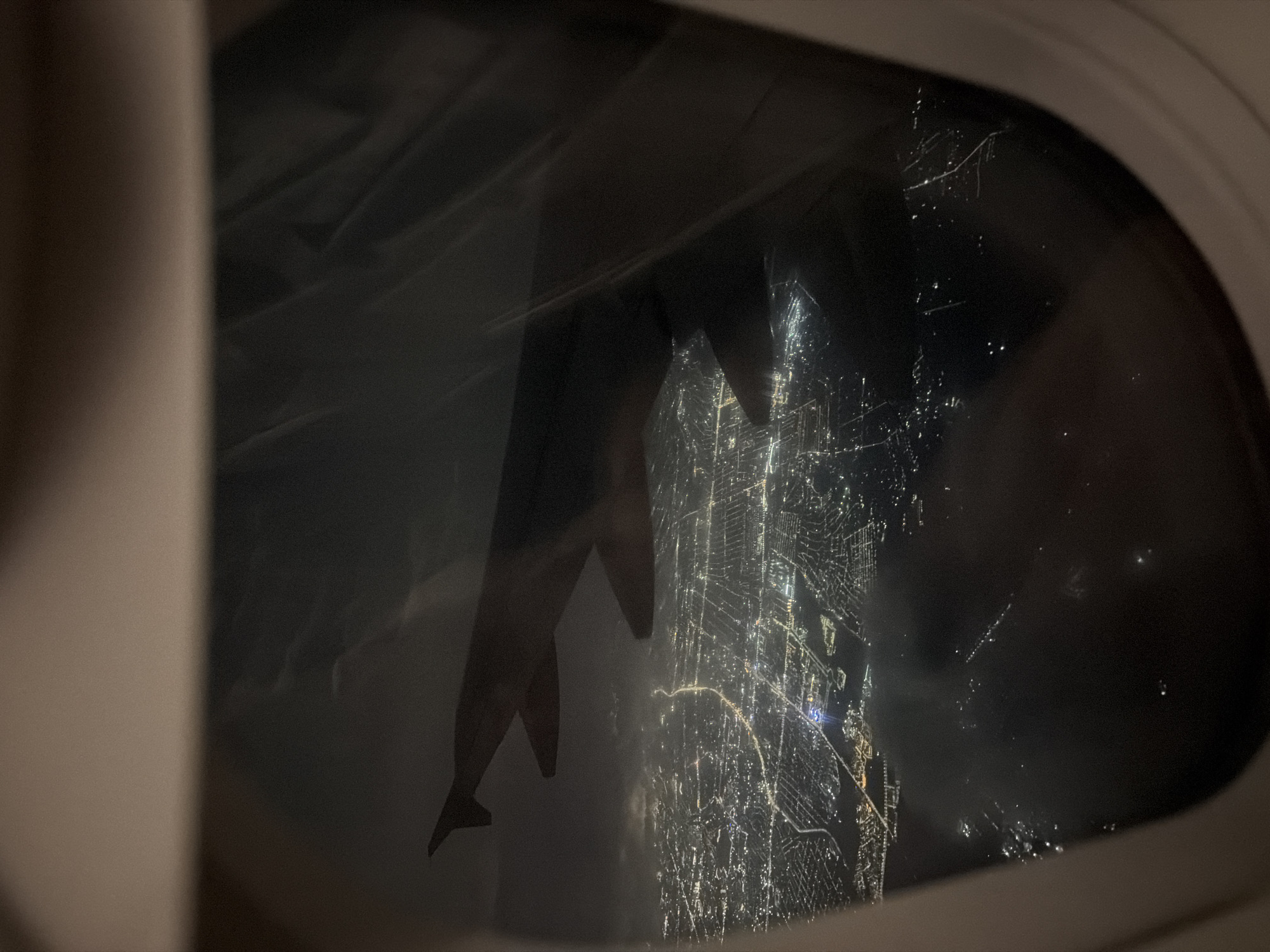 The image captures a view from an airplane window at night, looking down upon a sprawling city illuminated by countless lights. The wing of the aircraft is visible in the foreground, partially obscuring the cityscape. The city appears dense with buildings and a network of roads, crisscrossed by brighter lights that likely represent major thoroughfares. In the distance, the lights fade into a hazy glow, suggesting the vastness of the urban area. The overall impression is one of a vibrant, active city seen from a high vantage point. The image does not contain any information that would allow for the identification of the specific location within Köközek, Kazakhstan, or the presence of any individuals. The environment is consistent with an aerial view of a metropolitan area at night.