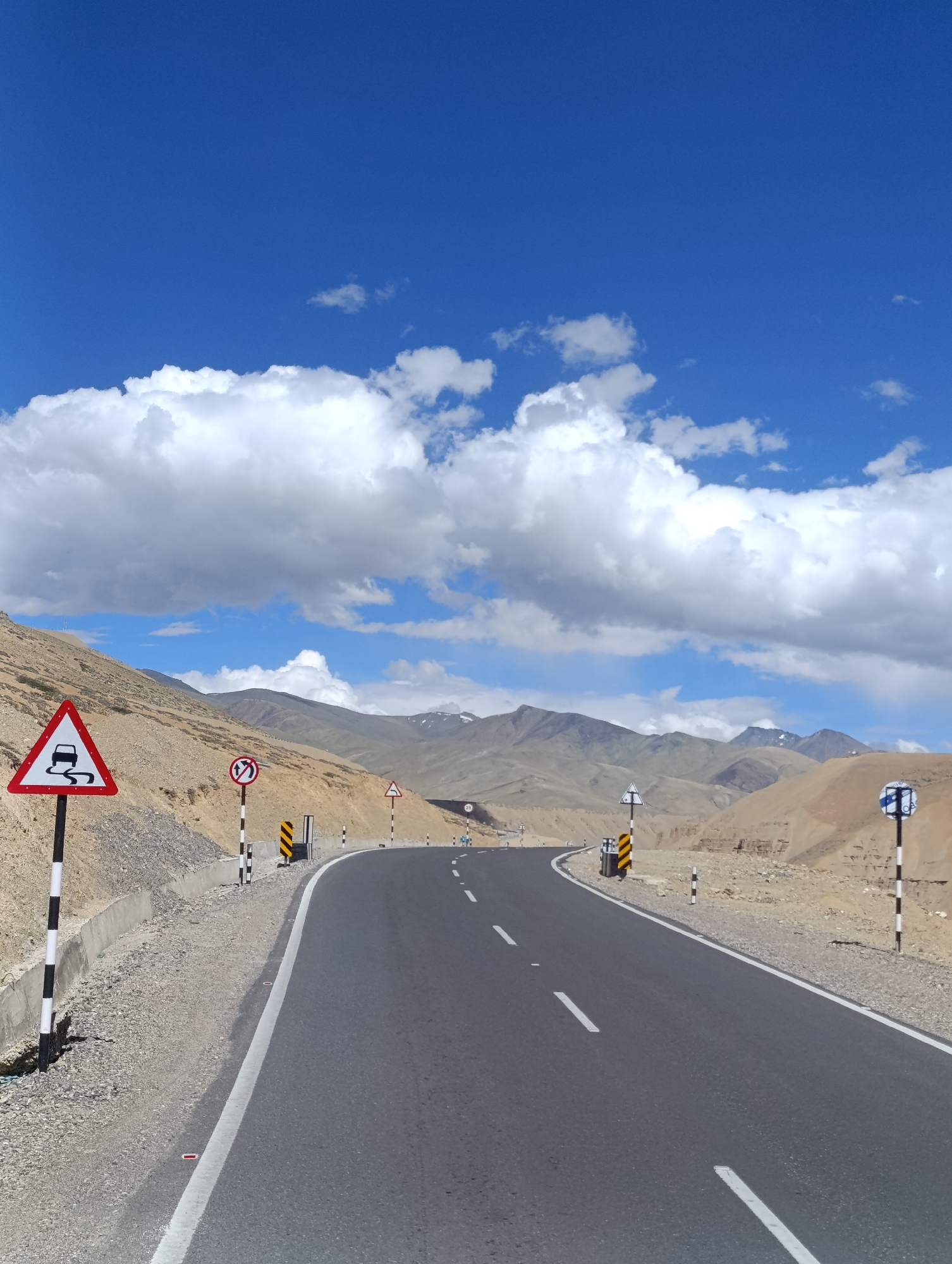 The image depicts a winding asphalt road through a mountainous, arid landscape under a bright blue sky with scattered white clouds. The road is bordered by a gravelly embankment on the left and barren, sandy hills on the right. Several road signs are visible: a triangular warning sign with a car skidding on a slippery surface, a circular sign with a diagonal red bar over an arrow indicating a sharp turn, and other smaller advisory signs. Mountain ranges with snow-capped peaks are visible in the distance. The scene is devoid of any people or vehicles.