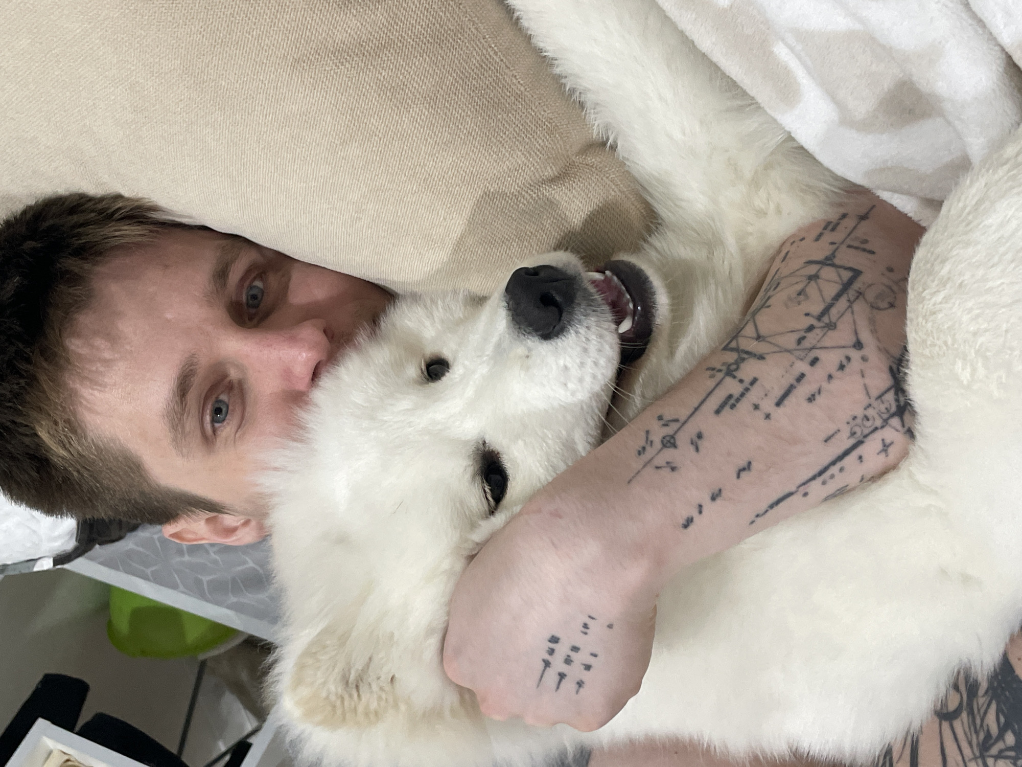 An eye-level shot captures a person and a white dog in a close, affectionate embrace, likely indoors on a bed or couch. The person, with light brown hair and blue eyes, is looking directly at the camera. Their head is partially resting against a light beige, textured pillow or cushion. The dog, which appears to be a large, fluffy white breed such as a Samoyed, is nestled tightly against the person, with its head turned towards the camera. Its mouth is slightly open, revealing its tongue and teeth, suggesting a playful or happy expression.
The person's left arm is draped around the dog, and this arm is extensively tattooed with intricate, geometric, and abstract patterns. Some designs resemble technical drawings or schematics, particularly on the forearm, while simpler, symbolic patterns are visible on the back of the hand. A soft, white, fluffy blanket or piece of fabric is visible covering part of the dog and around the person's tattooed arm, enhancing the cozy atmosphere.
In the background, on the far left, indistinct domestic items can be glimpsed, including a patterned surface and a splash of green from what might be a container. The overall impression is one of warmth, comfort, and a strong bond between the person and their pet. No specific time of day or outdoor weather can be determined, and there is no legible text visible within the frame.