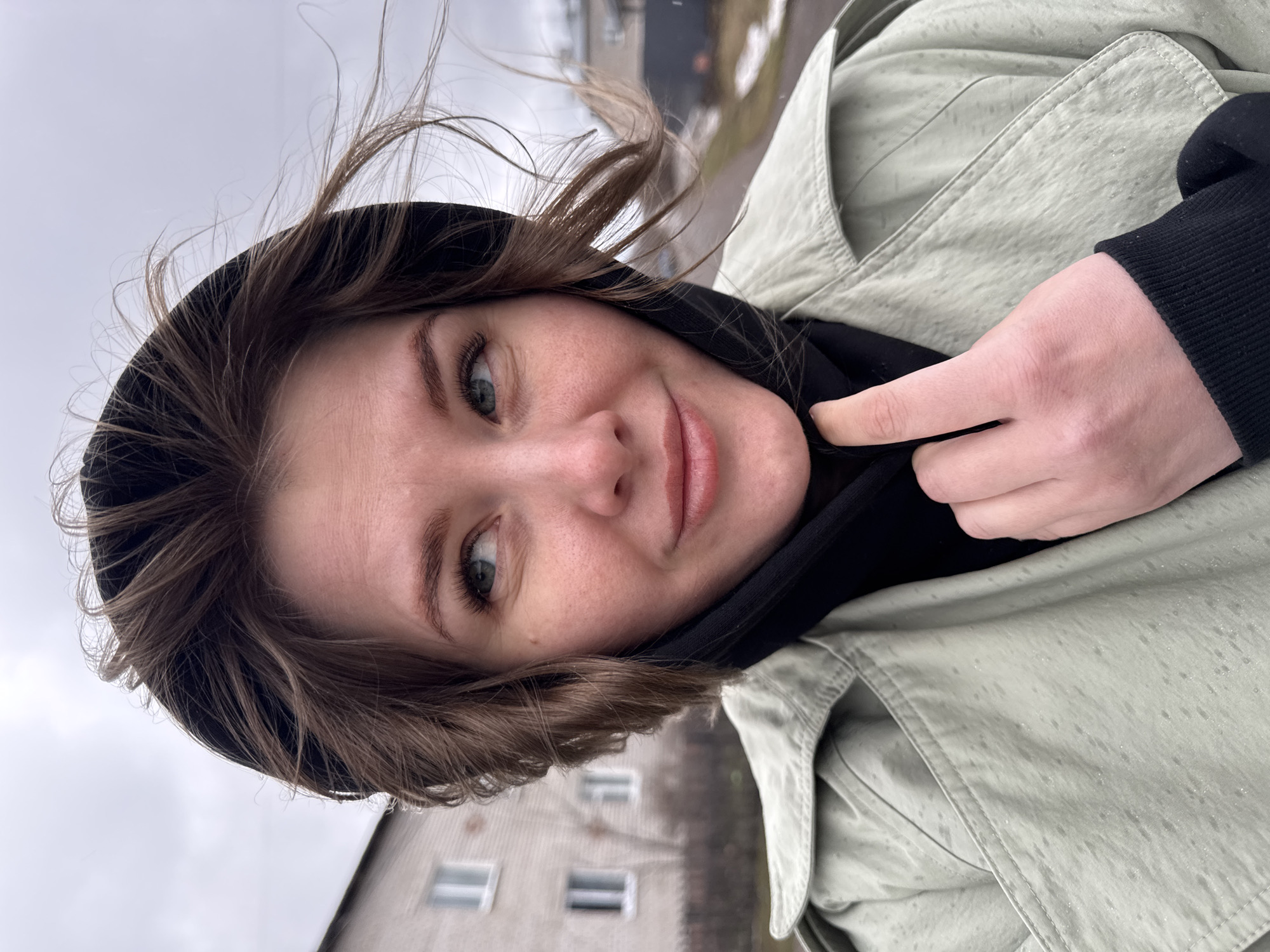The photograph is a selfie of a light-skinned young woman with light brown hair and blue eyes. Her hair is windblown and partially covered by a black hood. She is wearing a light green trench coat and a black hooded sweatshirt. She is pointing to her neck with her right index finger.
The background features a building with white-framed windows and an overcast sky. The image was taken in Горный, Russia. Rain or snow is present on the jacket.