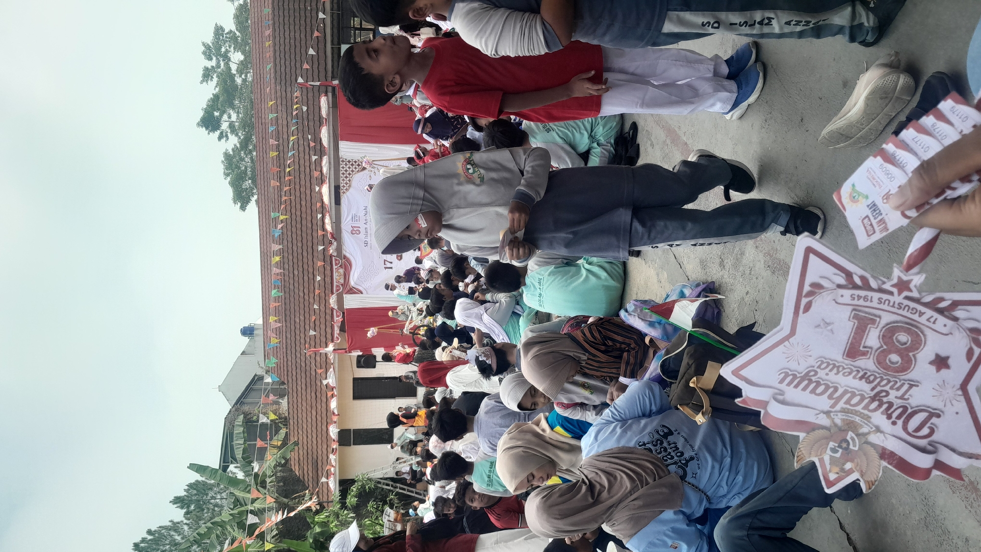 The image depicts an outdoor gathering, likely a school event, at SD Islam An-Nahl in Tasikmalaya, Indonesia. A large crowd of children and adults are seated on the ground, facing a stage decorated with red and white banners. Many children are wearing uniforms, some with hijabs. A young girl in a gray hijab stands in the foreground, holding a small item in her hand. A man's hand holds a red and white star-shaped decoration with the text "Dirgahayu Indonesia 18" and a stamp with the Garuda Pancasila symbol. Another hand holds several small paper tickets. The environment appears to be a schoolyard with buildings and trees visible in the background. The overall atmosphere suggests a festive occasion, possibly celebrating Indonesian Independence Day.