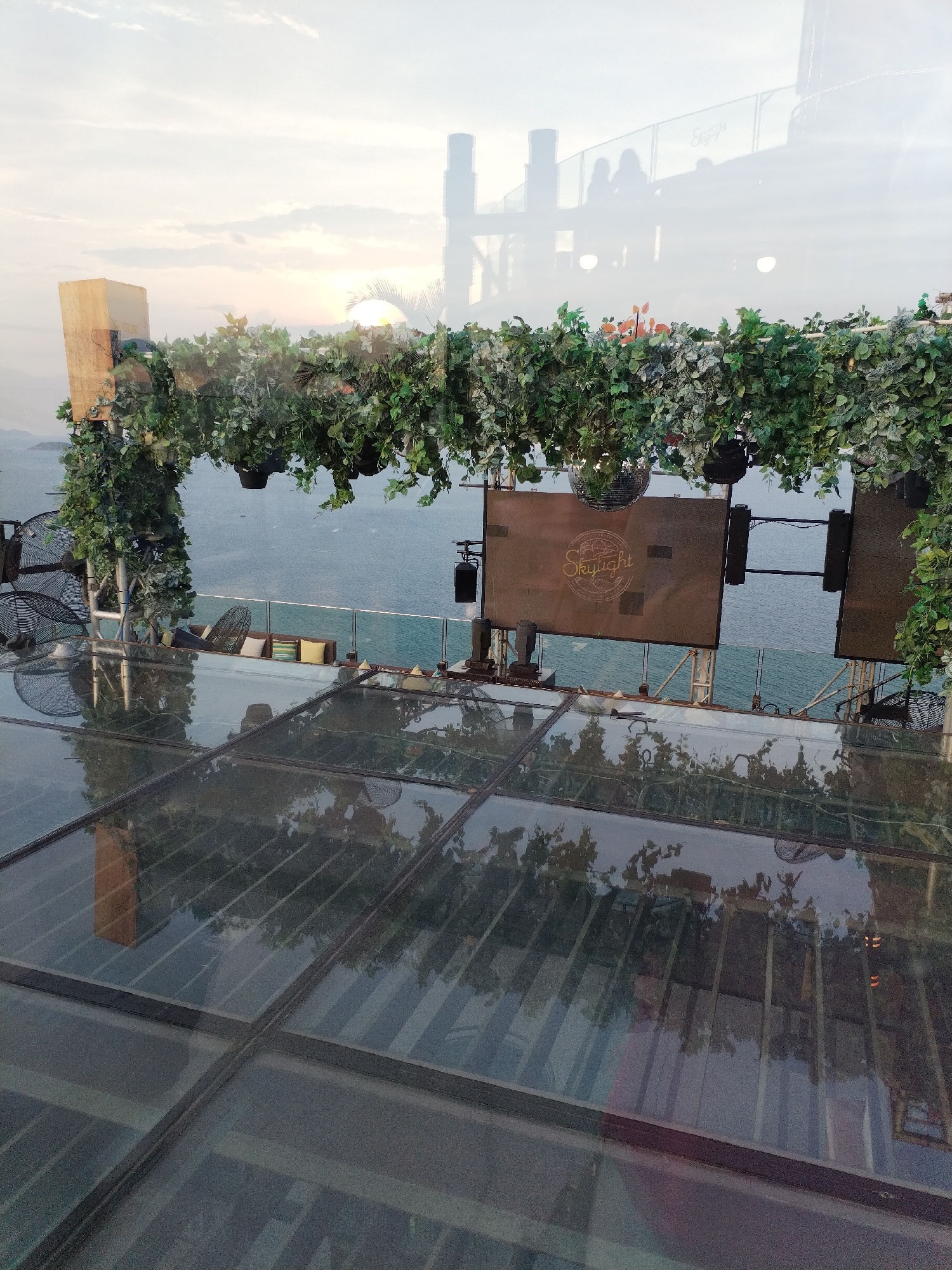 The image captures a rooftop scene in Nha Trang Ward, Vietnam, likely a bar or restaurant, with a glass floor that reflects the sky and surrounding structures. A canopy of artificial green foliage adorns the upper portion of the frame, framing a view of the ocean and a hazy sunset. Two large screens are visible, one displaying the word "Skylight." Silhouettes of people can be seen in the reflection on the glass, suggesting the presence of patrons enjoying the view. The overall atmosphere is one of a modern, scenic establishment overlooking the water, possibly at dusk.