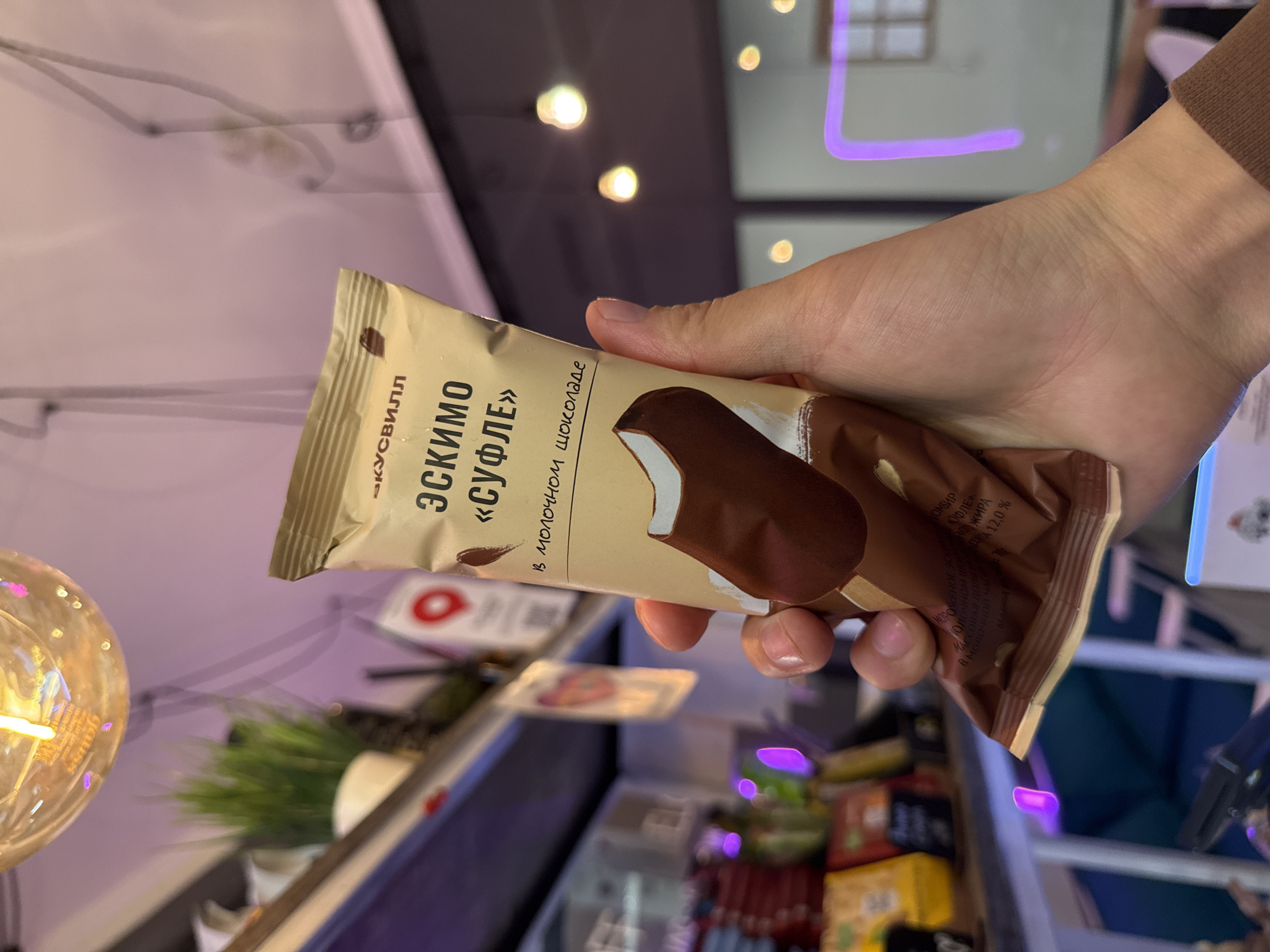 A person is holding a chocolate ice cream bar, labeled "Эскимо «Суфле»" (Eskimo "Soufflé"), in a tan wrapper. The wrapper features a picture of the ice cream bar covered in chocolate. Text on the wrapper, visible in Russian, includes "в молочном шоколаде" (in milk chocolate) and nutritional information. The scene appears to be indoors, possibly a shop or store, with shelves of products in the background, including some brightly colored packaging. A decorative light bulb with visible filaments is in the foreground, and hints of purple lighting suggest an artificial environment. The person's hand holding the ice cream is the primary subject. The lighting suggests it could be daytime or evening, with artificial light being prominent.