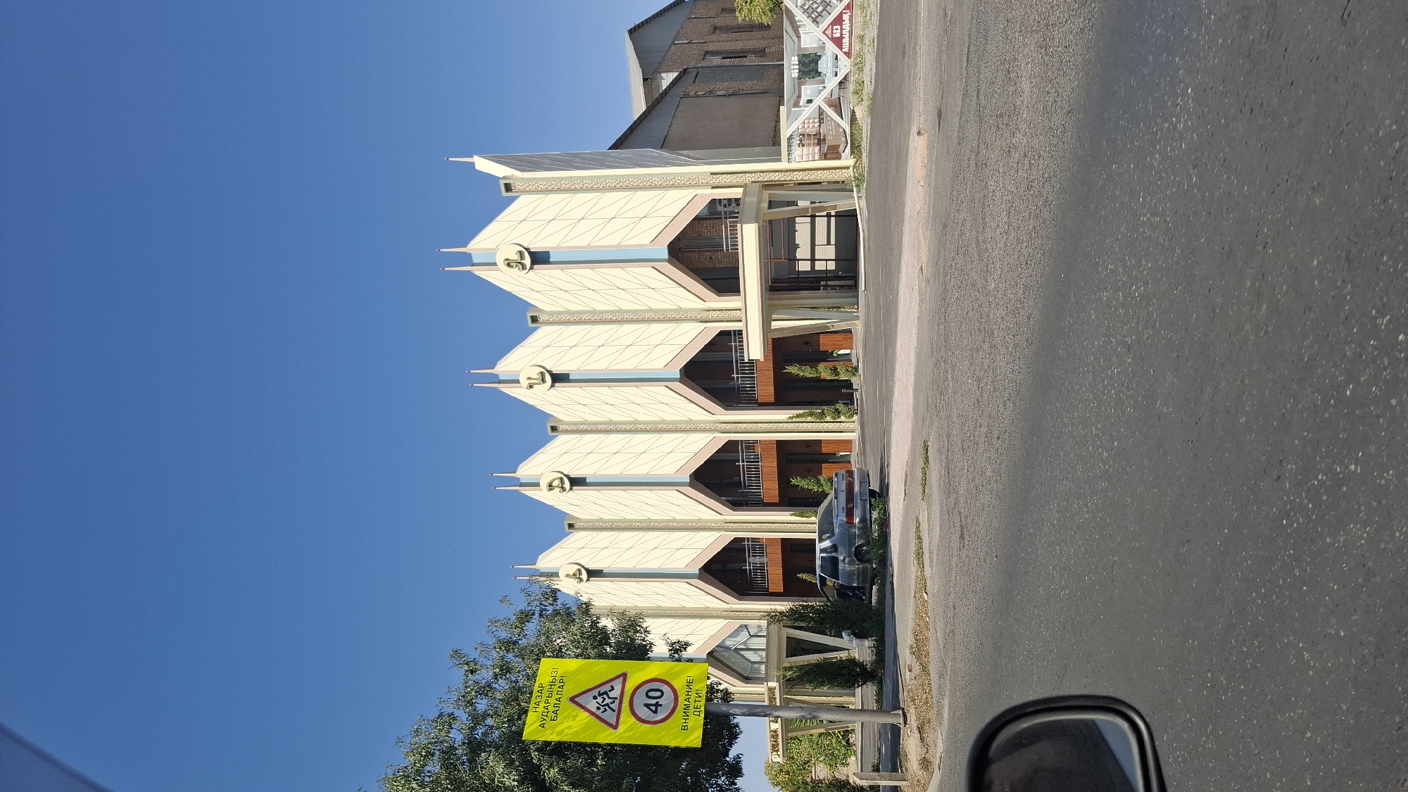 The image captures a street view in Shymkent, Kazakhstan, on a clear, sunny day. In the foreground, a portion of a car's side mirror is visible on the left, reflecting the sky and a tree. The road surface is asphalt. To the right of the mirror, the street stretches towards a building.
Dominating the midground is a distinctive building with a stylized, angular facade, featuring pointed roof elements and decorative arches. The building is light-colored, possibly cream or beige, with accents of blue and possibly pink or peach. A prominent yellow traffic sign with black text and symbols is positioned on the left side of the road in front of the building. This sign appears to be a warning sign, indicating a speed limit of 40 and a caution for children.
A silver sedan is parked on the side of the road in front of the building. Several small, conical trees are planted along the building's facade and in front of the car. In the background, another building, appearing more industrial and utilitarian with exposed brickwork, is visible to the right of the main structure. The sky is a vibrant, cloudless blue. The overall impression is of a commercial or public building on a street in Shymkent.