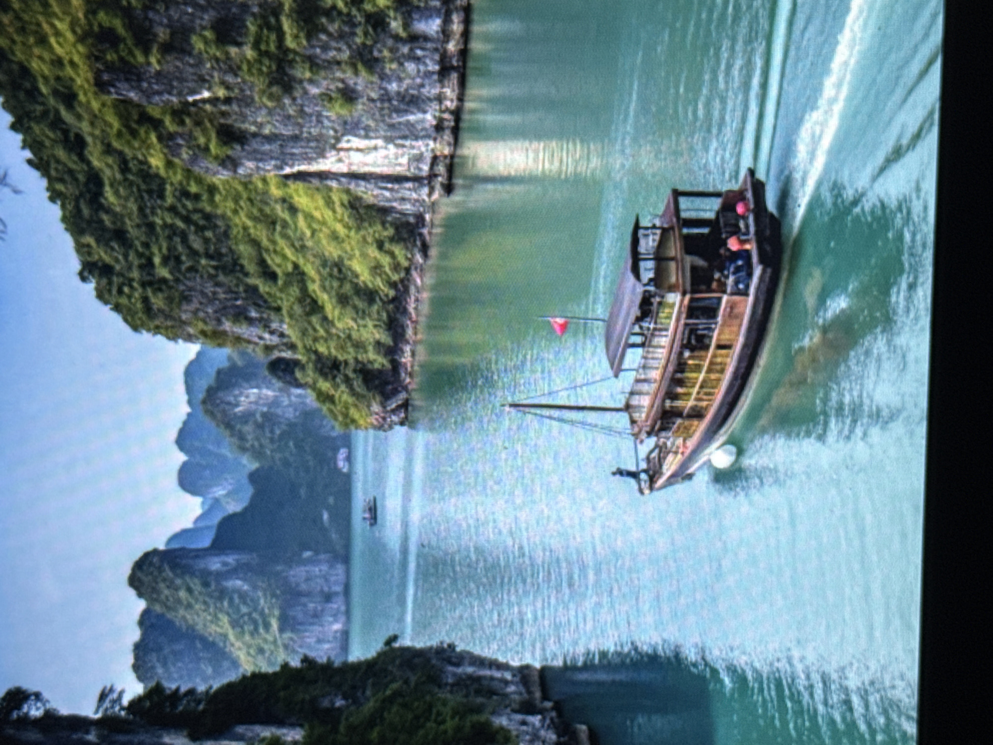 A long, traditional wooden boat with multiple decks and a red flag is sailing on a calm, turquoise body of water. The boat has people visible on its decks and appears to be moving from left to right, leaving a wake. The surrounding environment consists of steep, lush green islands with rocky cliffs rising from the water. The sky is a clear, pale blue. The overall scene depicts a serene landscape, likely a bay or inlet known for its natural beauty and boat tours.