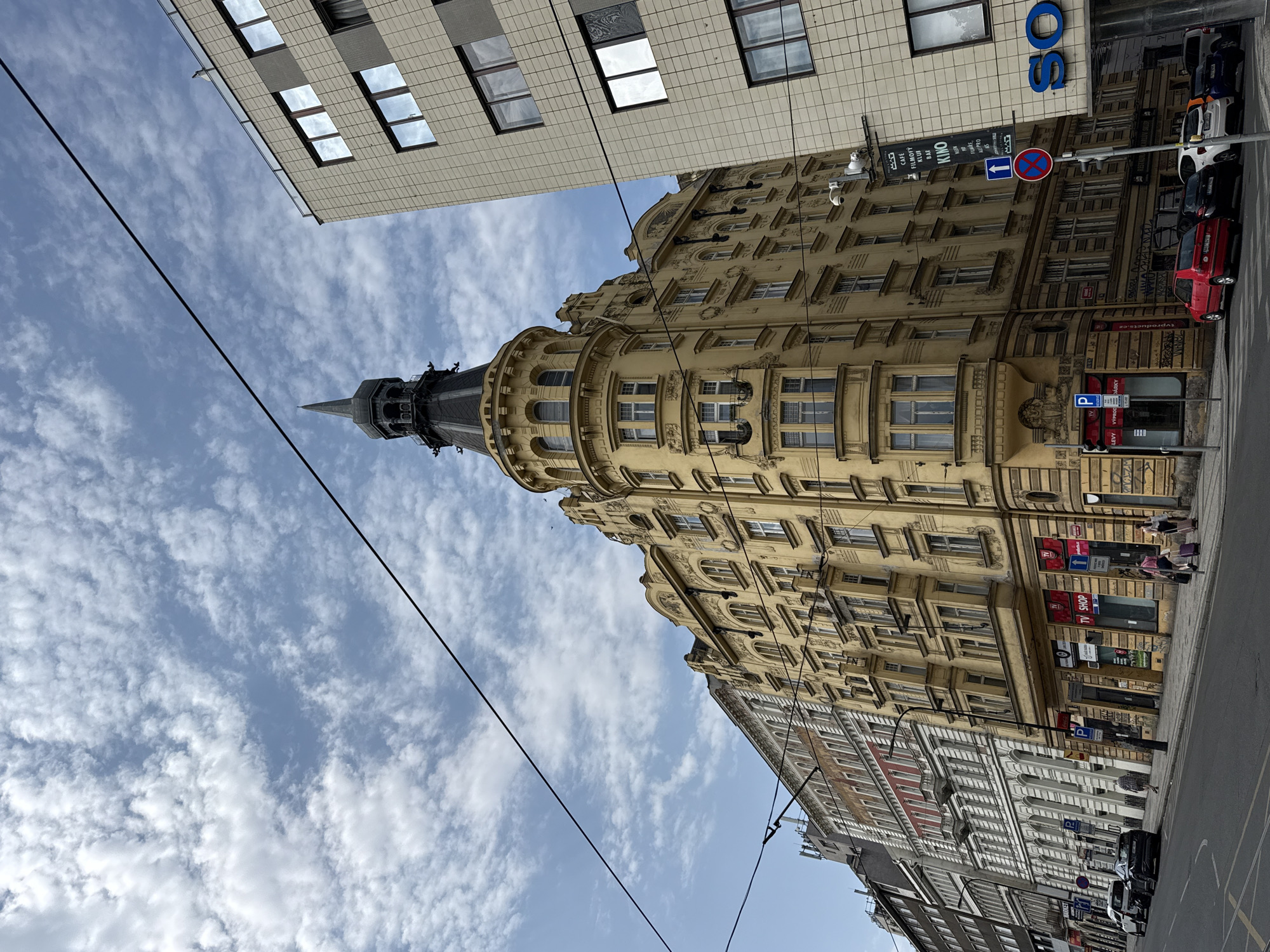 This is a street-level, eye-level, medium shot of a street scene in Prague, Czechia. The scene is dominated by historical buildings and a modern office building on the right. A prominent yellow, ornate building with a tower and a spire stands in the center left of the frame. This building features multiple floors with numerous windows and decorative architectural details. To the right of this historical building is a modern, light-colored building with a grid-like facade and large windows. The sky above is blue with scattered white clouds. Several power lines are visible stretching across the frame.
On the street level, there are parked cars and a sidewalk. Several people are walking on the sidewalk, some with luggage. A traffic light and a blue "P" parking sign are visible near the corner. A black banner with white text, advertising "KINO" and other services, hangs from a pole on the modern building. Below the banner is a circular red and blue traffic sign. The buildings on the far left of the frame appear to be older and have a different architectural style than the central yellow building. The overall impression is an urban environment where historical and modern architecture coexist.